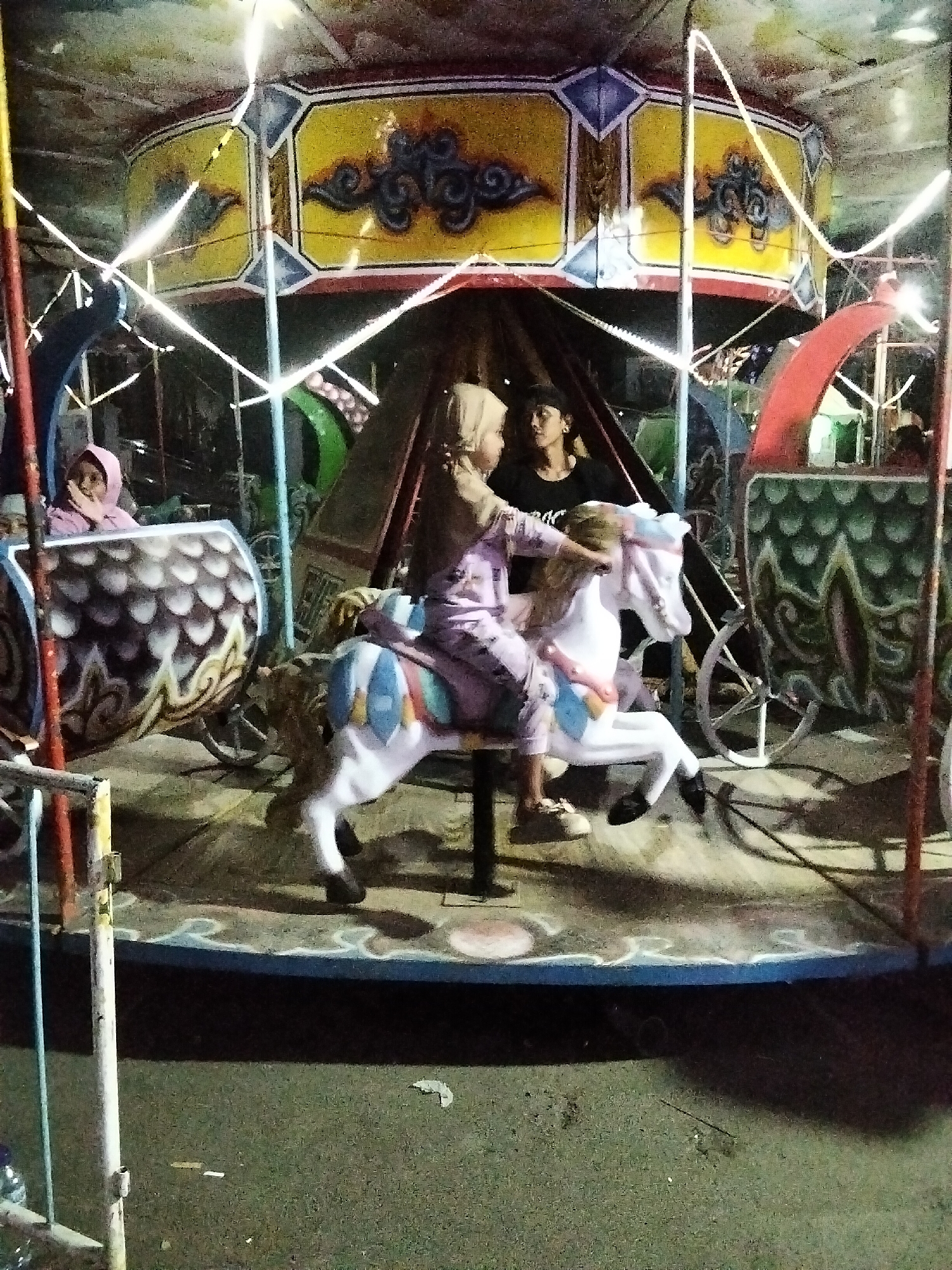 The image captures a carousel ride at night in Cisaat, Indonesia. The carousel features an ornate, brightly painted top with yellow, blue, and red accents, illuminated by strings of lights. A white fiberglass horse, adorned with blue, white, and red saddle details, is occupied by two young girls wearing headscarves. The girl in front is wearing a light purple outfit and is holding onto the horse's reins. The girl behind her, also in a light purple outfit, has her hand on the front girl's shoulder. To the left of the horse is a decorative carriage with a patterned exterior resembling scales, and to the right, another similar carriage is partially visible. In the background, a person with dark hair and a dark shirt is looking towards the girls. The ground around the carousel appears to be a mix of concrete and dirt, with scattered debris. The overall scene suggests a festive atmosphere at a local amusement or night market.