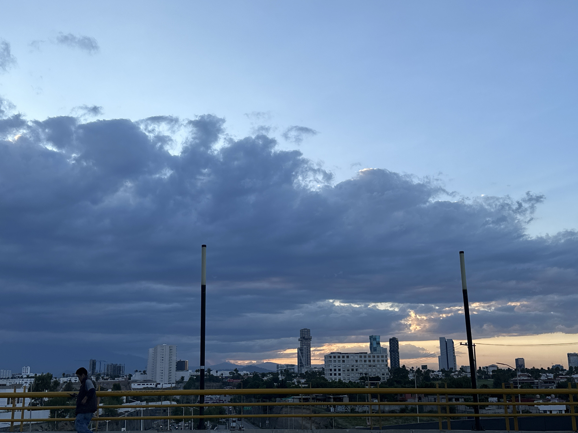 The image shows a cityscape at dusk under a cloudy sky. In the foreground, a yellow railing stretches across the width of the frame, suggesting a bridge or elevated walkway. Two tall, black and white poles stand upright along the railing.
To the left of the frame, a person wearing a mask is walking along the walkway. The person is dressed in casual clothing.
In the background, a row of buildings of varying heights is visible, interspersed with trees. The sky is a gradient of blues and grays, with patches of orange light peeking through the clouds near the horizon. The location appears to be a city, but the specific location is unknown.