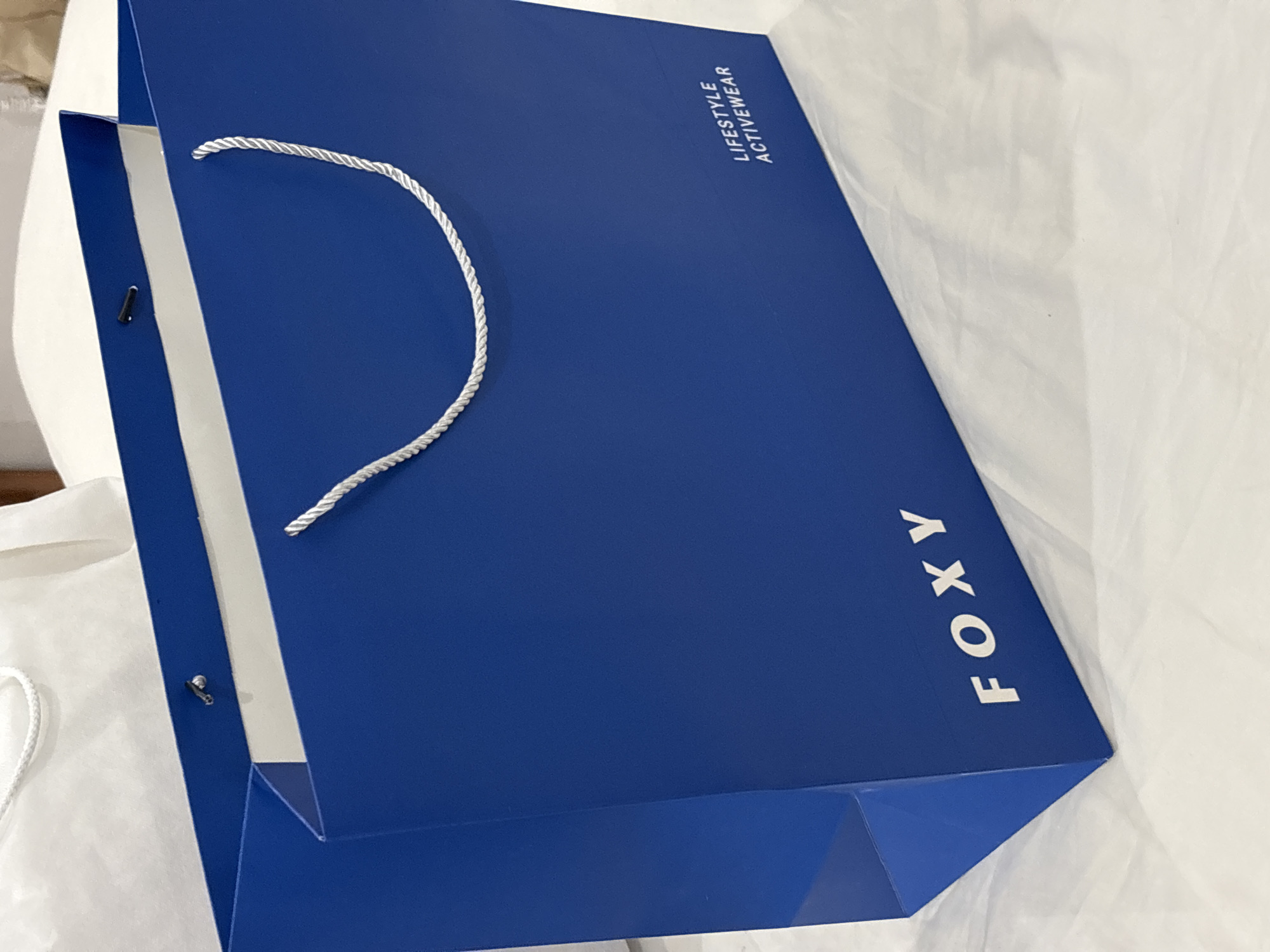 A close-up, angled shot focuses on a blue shopping bag with white rope handles. The bag, bearing the word "FOXY" printed vertically in white, also features the phrase "LIFESTYLE ACTIVEWEAR" in white, stacked text. The bag is situated on a white, slightly rumpled surface. Part of a white shopping bag is visible to the left of the blue one. The location is Kuta Selatan, Indonesia.