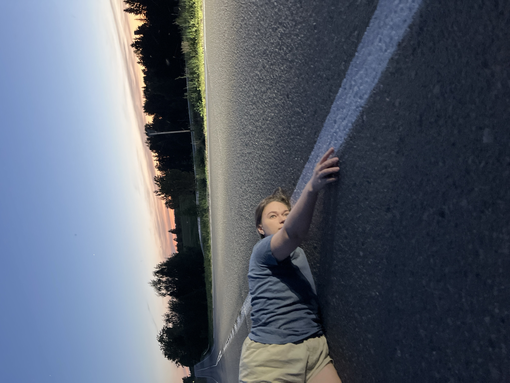 A medium shot shows a young person lying on a paved road in a rural area near Ольховка, Russia. They wear a blue t-shirt and light khaki shorts, extending their right arm to touch a white line painted on the asphalt. The sky behind the trees suggests it is dusk or dawn.