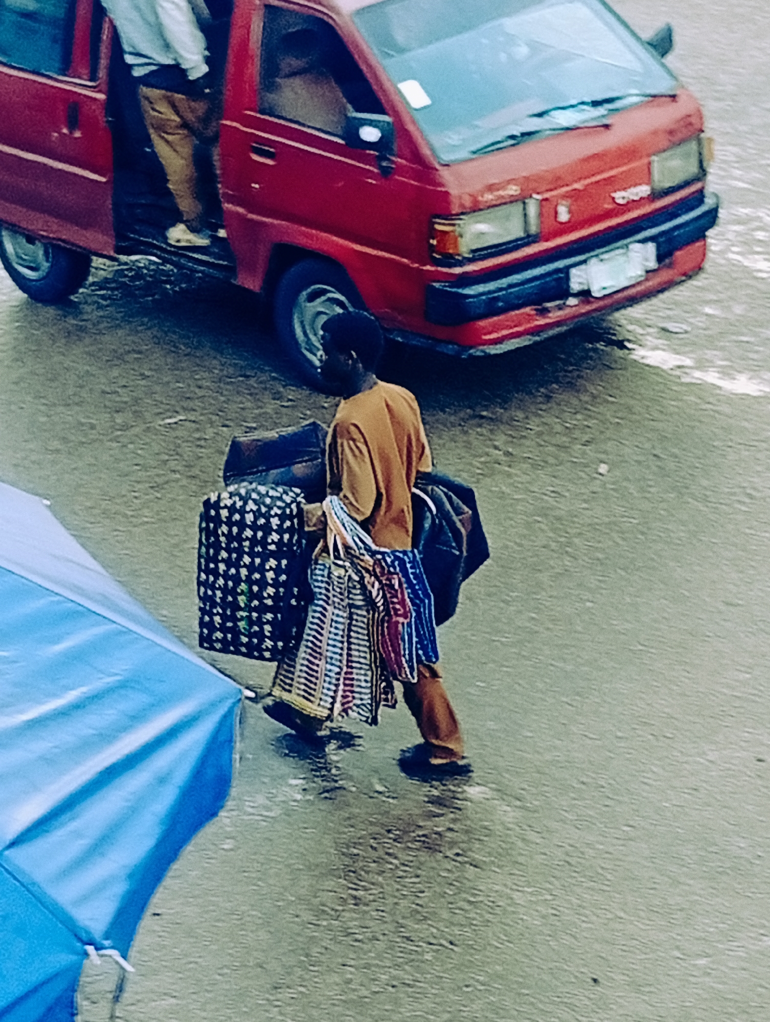 A person is walking across a wet, paved surface, carrying several bags. The person is wearing a brown shirt and brown pants, and appears to be a male. He is carrying a dark bag over his left shoulder, and several other items, including a patterned cloth bag and what looks like more fabrics or textiles, in his hands. To the left, a large blue umbrella is partially visible, also suggesting wet weather. In the background, a red van with its door open is parked. The van has a black bumper and appears to be a Toyota. The scene is set in Bukuru, Nigeria, and the wet ground indicates it is likely raining or has recently rained. The time of day is indeterminate, but the lighting suggests it could be daytime. The overall mood is that of a person engaged in transit with their belongings, possibly dealing with the inconvenience of wet weather. No visible text is clearly legible on the van or other elements in the image.