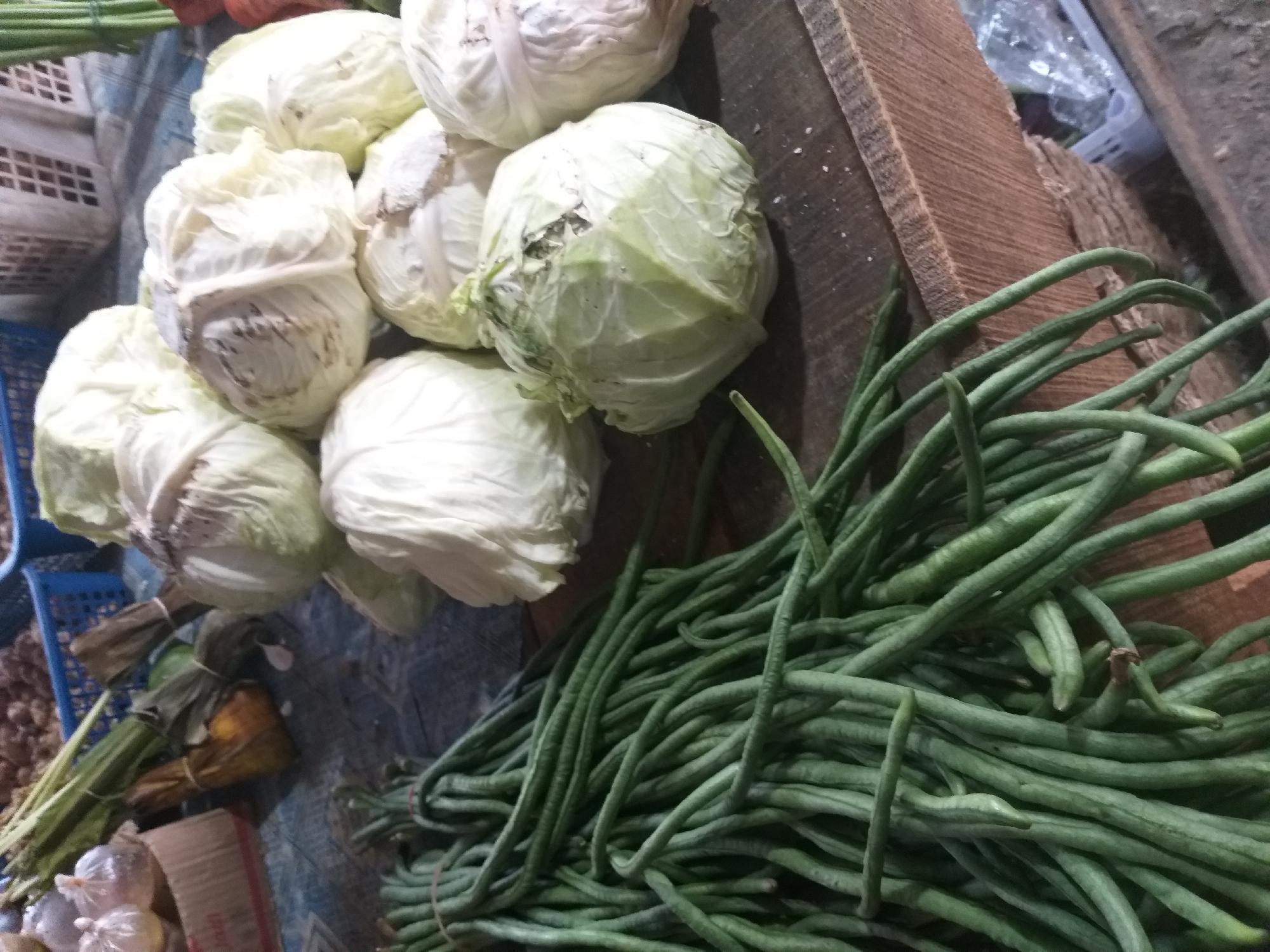 A still life image captures a selection of fresh produce displayed on a wooden surface, likely in a market stall within Air Jamban, Indonesia. In the foreground, a large bundle of long, green beans, tied with string, lies spread across the wooden table. To the right, several heads of pale green cabbage are piled, some appearing slightly wilted or with minor imperfections on their outer leaves. The background is blurred but suggests a market setting with other items and possibly baskets. The lighting is diffused, and the overall impression is of a humble, local market scene.