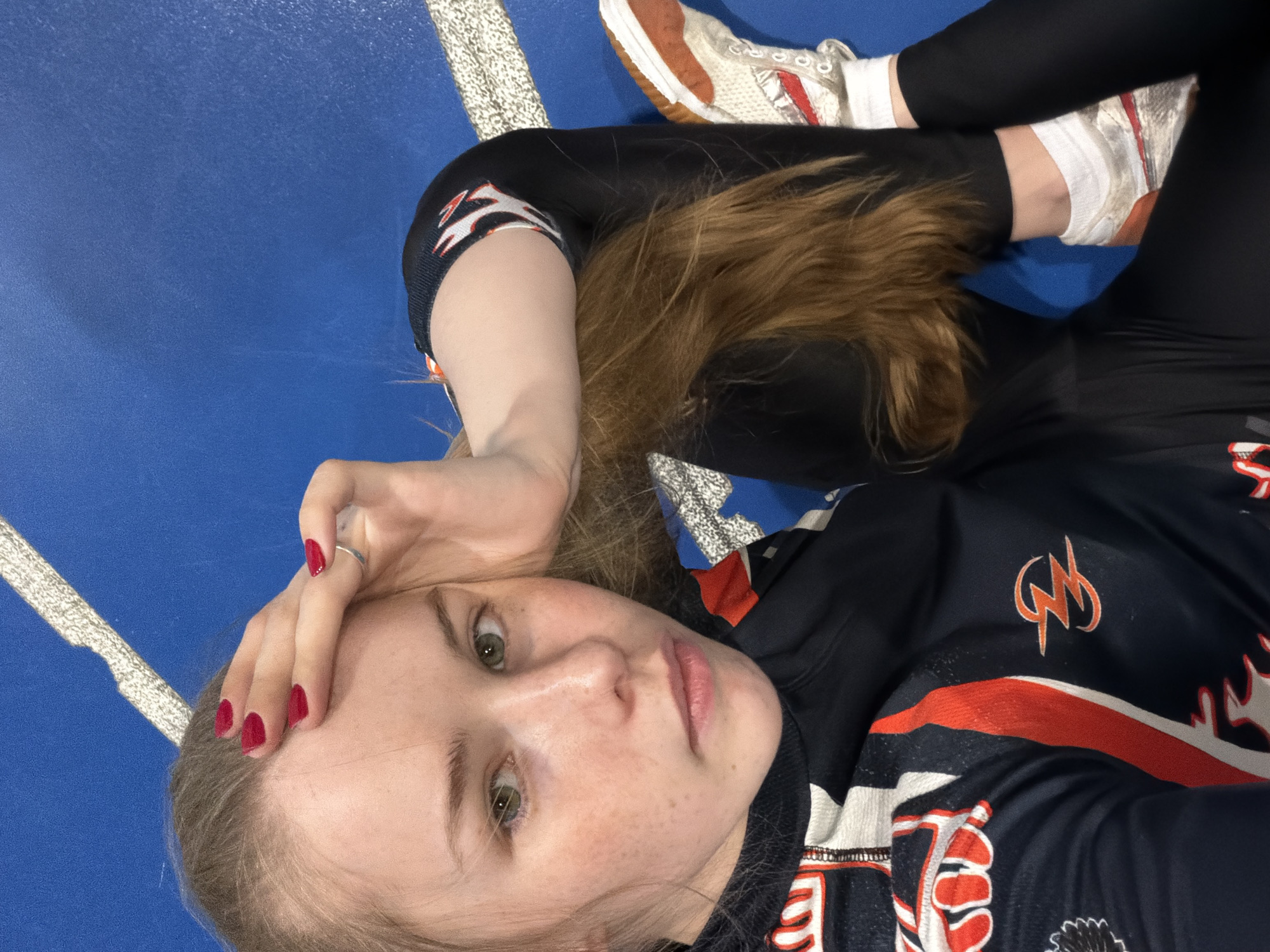 A young person with light skin, freckles, and green eyes is lying on a blue surface. The person is wearing black leggings, a black shirt with orange and white markings, and white sneakers with brown soles and red stripes. The person's hair is light brown. The person's left hand is near their face, and the fingernails are painted red. The location in the city of Berezniki, Russia, is not discernable from the image.