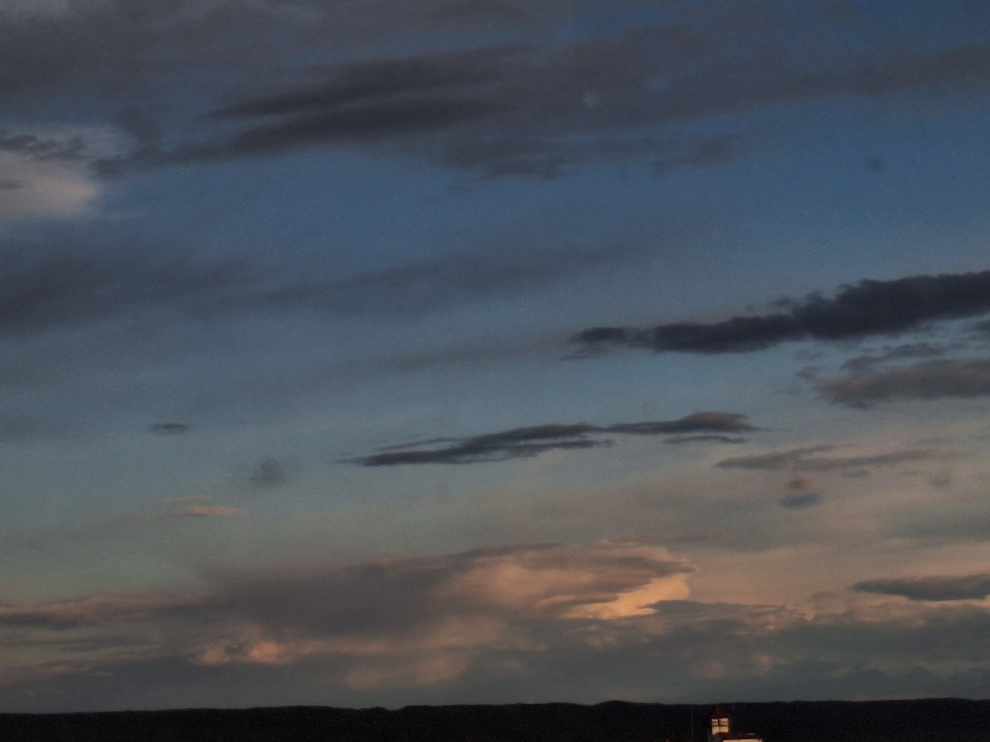 The image depicts a twilight sky over what appears to be a dark, distant landscape. The sky is filled with varied cloud formations, ranging in color from dark gray and blue to lighter shades of pink and orange where the sun is setting. At the bottom of the frame, a dark silhouette suggests a landmass or a building, and a lit structure is partially visible. Without further details it is hard to identify objects or the location. It is not possible to say anything about the city of Anipemza, Armenia.