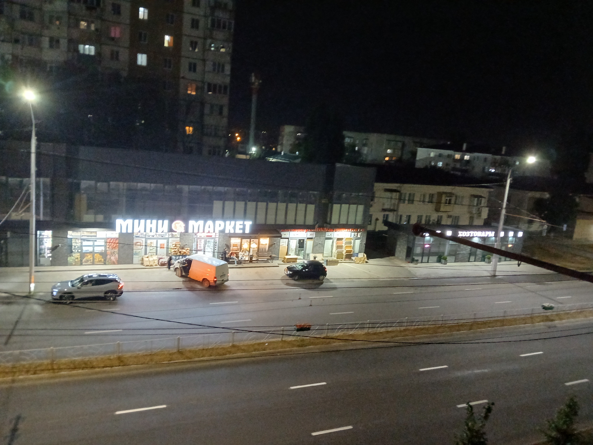 The image shows a street scene at night in Cherkessk, Russia. A multi-lane road with white lane markings runs horizontally across the foreground. To the left of the road, a brightly lit storefront with the sign "МИНИ МАРКЕТ" (Mini Market) is visible. In front of the market, several vehicles are parked or driving. A silver SUV is moving on the road, and a white van with an open orange cargo door is parked near the market. Another dark-colored car is also visible on the road. Across the street from the mini-market, another storefront with illuminated windows is present, possibly a department store or another retail establishment, with signage that appears to read "ХОЗТОВАРЫ" (Household Goods). In the background, apartment buildings are illuminated with lights from windows, indicating residential activity. A street lamp is also visible on the left side of the image. The overall scene depicts a commercial and residential area at night.