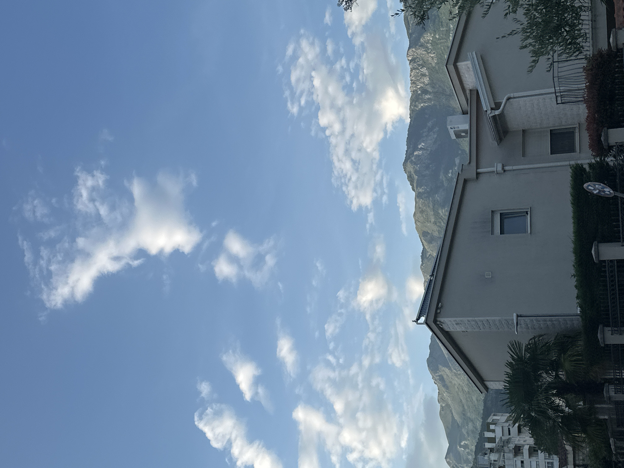 The image depicts a daytime scene in Budva, Montenegro. The sky is predominantly blue with scattered, puffy white clouds. A mountain range forms a backdrop to the right, partially obscured by the buildings. Two light-colored buildings are visible. The taller one on the right has an air conditioning unit mounted on the wall. A portion of a street lamp and some trees are also visible in front of it. Further down and to the left is a second, smaller building, partially obscured by a palm tree. In the bottom left, a building or construction site is visible.