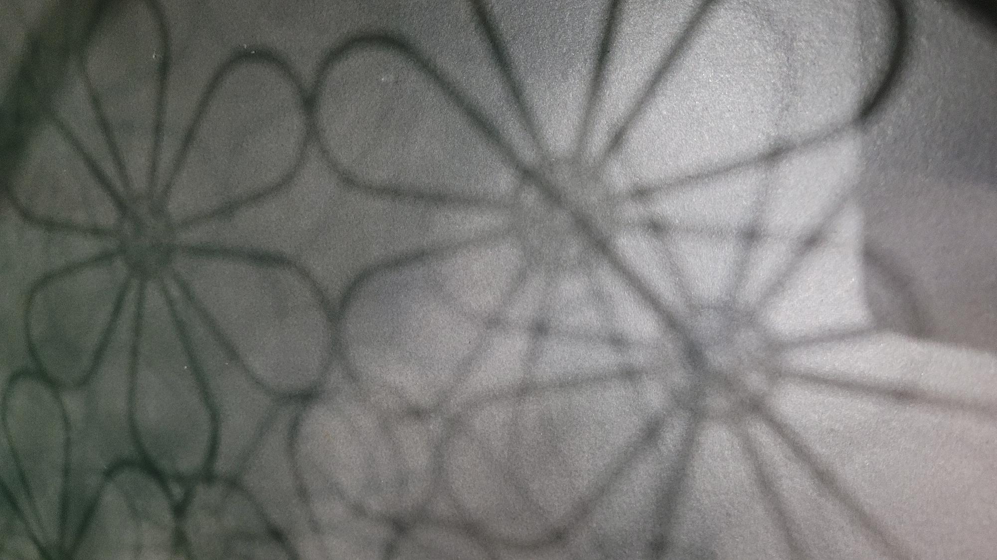 This is a close-up, angled photograph of shadows cast on a textured, grey surface. The shadows form abstract, stylized floral or starburst patterns, with thin, dark lines radiating from central points and connecting to curved petal-like shapes. The lighting appears to be coming from above and to the side, creating soft edges and variations in shadow density. The surface itself has a granular texture, reminiscent of concrete or rough paper. There are no visible people, objects, or environmental features beyond the immediate surface and the shadows themselves. The location is unknown.