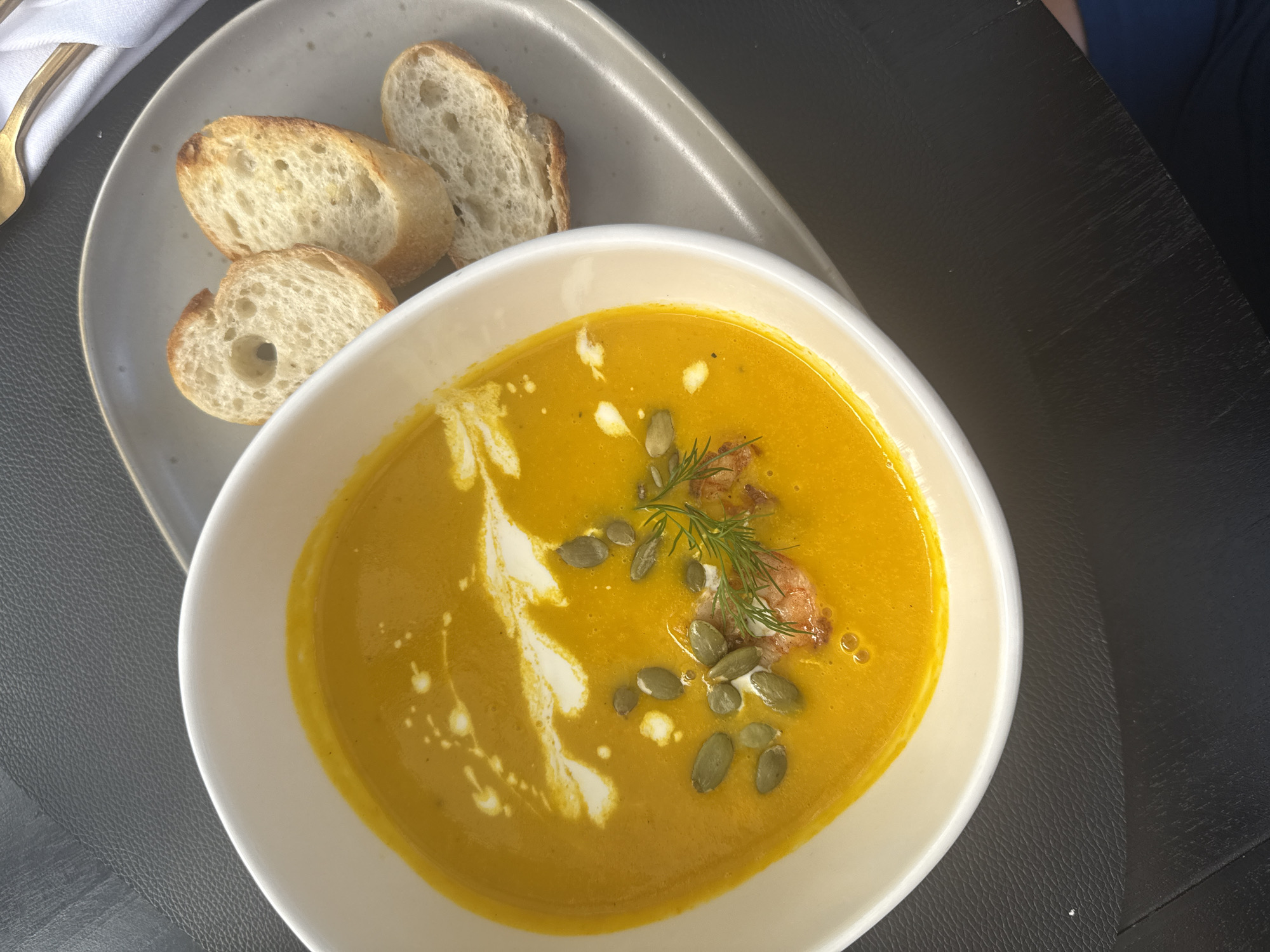 A top-down shot shows a bowl of bright orange soup, likely squash or pumpkin, garnished with a swirl of cream, scattered green pumpkin seeds, and a sprig of dill, accompanied by three slices of toasted baguette on a gray plate. The scene takes place on a dark, textured surface, perhaps a table at a restaurant or cafe. The location is not discernible from the image. No people are fully visible, although a hint of a blue-clad arm is present on the top right. The primary action is the presentation of food. The scene appears to be indoors, based on the lighting and the nature of the table setting.