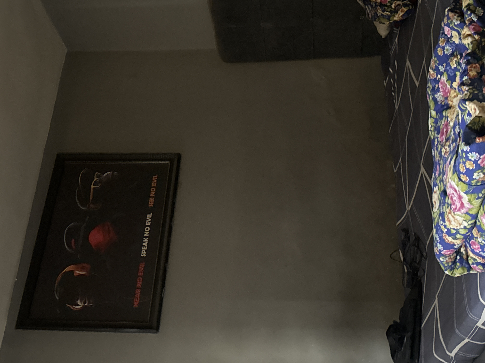 The image displays a dimly lit indoor room, likely a bedroom, viewed from an elevated perspective looking down into a corner. The overall ambiance is subdued due to the low lighting, suggesting either evening, night, or a room with restricted natural light.
On the left wall, a framed picture is a focal point. The artwork within the black frame features three stylized, dark figures, characteristic of the "three wise monkeys" motif. Above each figure, text is clearly visible: "HEAR NO EVIL" on the left, "SPEAK NO EVIL" in the center, and "SEE NO EVIL" on the right. Some details on the figures, such as eyewear and head coverings, appear in reddish and lighter tones against the dark background.
The right side of the image is dominated by a bed. It is covered with a dark grey or blue duvet featuring a geometric pattern of white intersecting lines. Partially draped over this is a colorful, floral patterned blanket or comforter, primarily blue with prominent pink, green, and white flowers. Below the floral blanket and towards the foot of the bed, some dark fabric or clothing items are visible. In the upper right corner, a dark, rectangular object, possibly a piece of furniture or a mat, rests on the floor. The walls of the room are a plain, light greyish-brown color.
No specific activity or event is taking place in the image, showing an unoccupied space. The scene is an interior room located in Kurunduma, Nigeria.