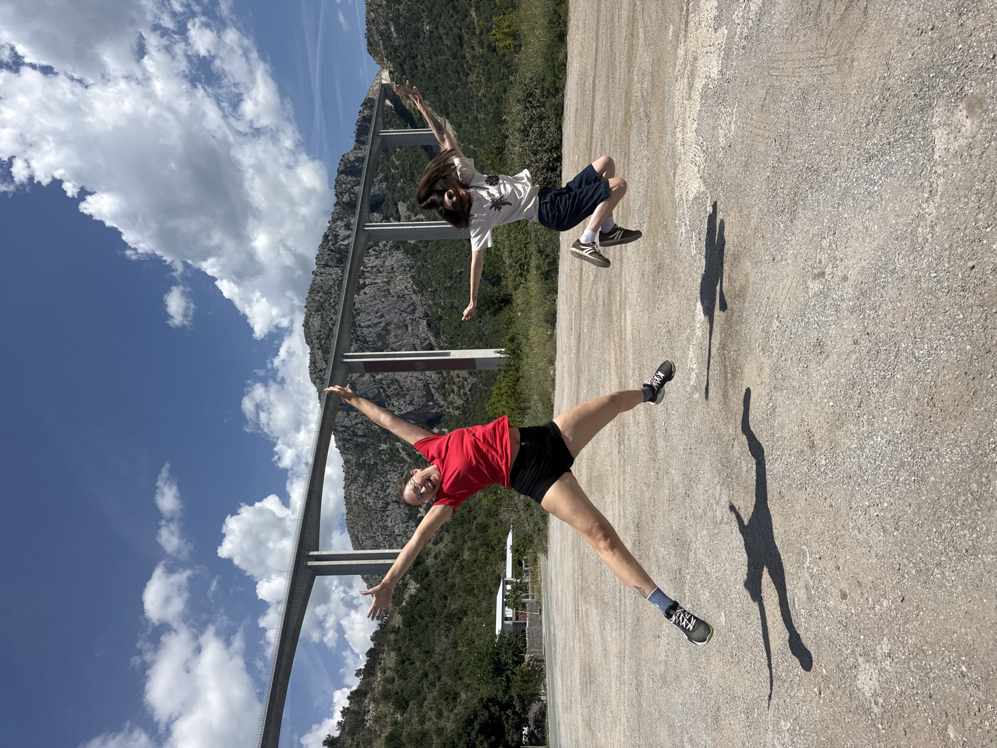 The image depicts two individuals in a mountainous region, with a large bridge structure in the background. The woman in the foreground, wearing a red shirt and black shorts, is striking a pose with outstretched arms and legs, appearing to be jumping or suspended in mid-air. The second person, wearing a white shirt and blue shorts, is also in a jumping pose, situated higher in the frame. The ground appears to be a gravel surface. In the background is the Mala Rijeka viaduct in the city of Bioče, Montenegro. The sky is visible, with clouds present. There are shadows cast upon the gravel surface.