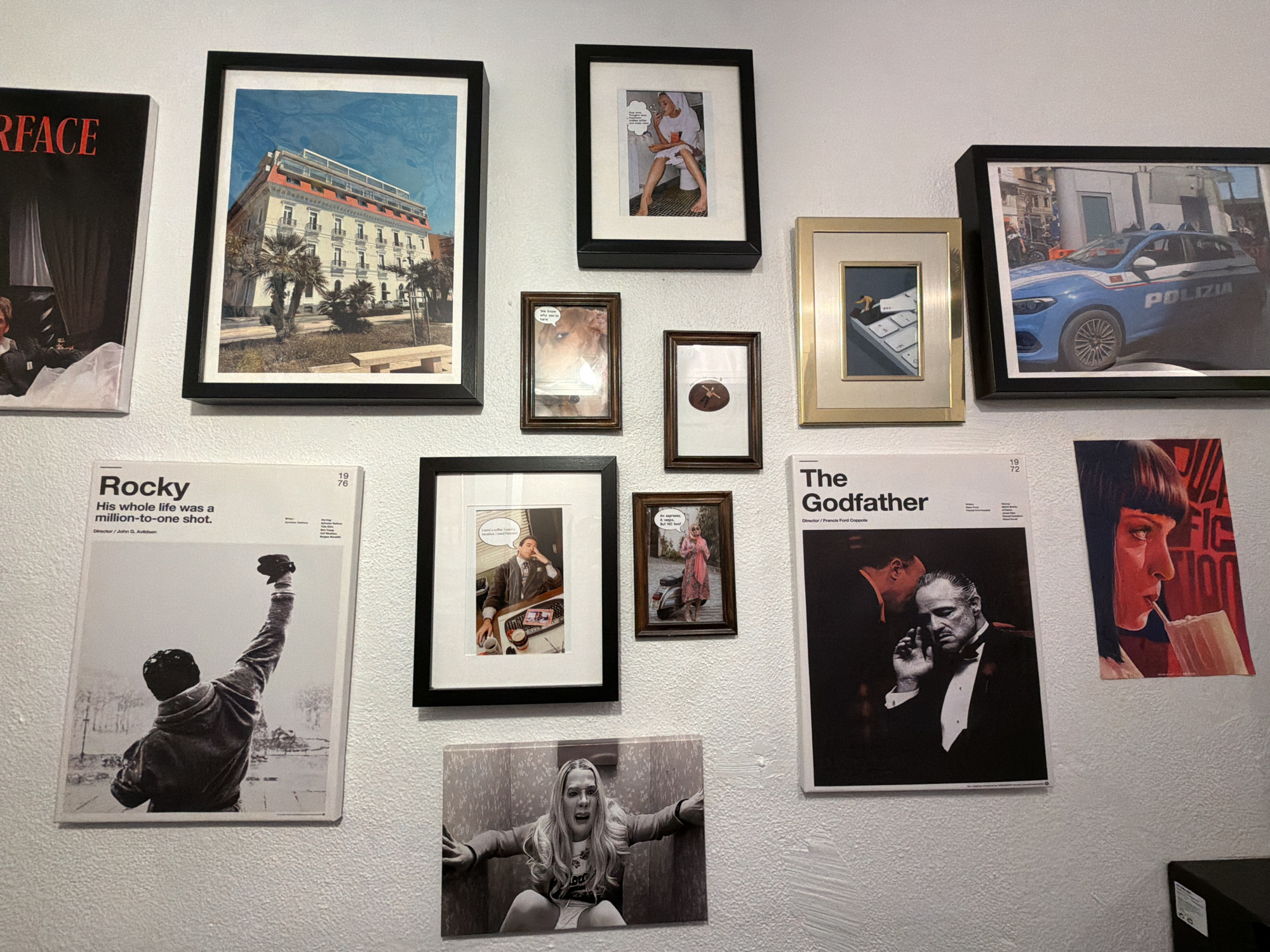 The image shows a wall covered with various framed photographs and movie posters. The posters include "Scarface," "Rocky," and "The Godfather." Framed photos depict a building with palm trees, a person in a bathtub, a dog with a thought bubble, a person rowing a boat, and a police car. Another photo shows a person at a desk with a thought bubble, and another shows a person in a dress standing next to a scooter. A final photo shows a person trapped in an elevator. The environment appears to be an indoor wall, likely in a residence or business, with the wall painted white. The subject matter is popular culture and film references. The location is not Montreal, Canada.