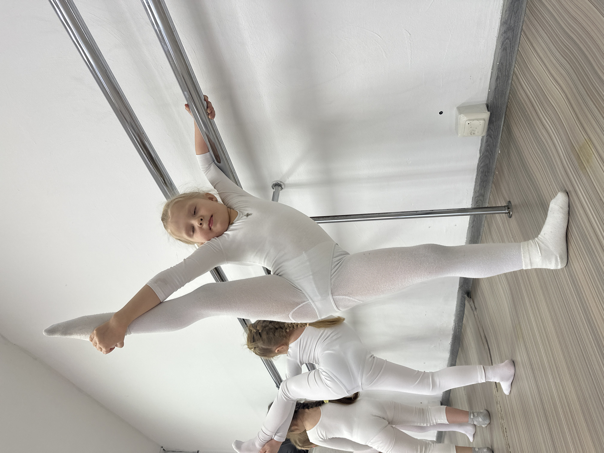 The image captures an indoor scene, likely a gymnastics or dance studio, with three young girls dressed in white performing acrobatic or stretching exercises. The main subject is a young girl with blonde hair, centered in the upper half of the frame, executing a straddle split while holding onto two parallel, chrome-plated metal bars. She is wearing a white long-sleeved leotard or bodysuit, white tights, and white socks, and looks towards the camera with a slight smile.
Below her, two other girls in identical white attire are visible, also engaged in stretching or split poses, positioned as if on lower bars or the floor. One of these girls has braided hair, differentiating her from the main subject.
The setting features white walls and a light-colored wooden or laminate floor. The metal bars are prominent, extending across the frame, suggesting a training facility. On the upper right wall, a white electrical outlet or switch box is visible, with faint text that appears to read "GLI" or "GCL". The image is taken from a low angle and rotated clockwise, creating a dynamic perspective of the girls' flexible poses. The overall atmosphere suggests a focused training session.