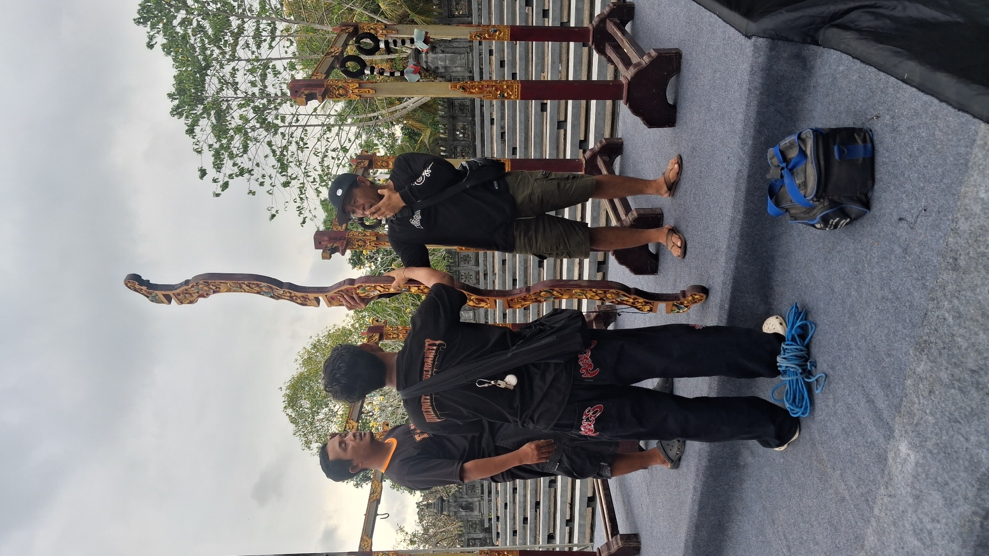Three individuals are gathered on a raised, gray platform, likely a stage, in Negara, Indonesia. They are positioned around an ornate, carved wooden structure, possibly part of a musical instrument or decorative element. The sky above is overcast.
On the left, a man wearing a black t-shirt with an orange collar and black shorts looks towards the center. In the middle, a person with their back to the camera, wearing a black t-shirt with "UNLIMITED SOLIDARITY" printed on it in white, and black pants with red lettering, holds the wooden structure. To the right, a man in a black long-sleeved shirt, olive-green cargo shorts, and a black baseball cap is speaking and gesturing with his right hand near his mouth. He is wearing sandals.
On the platform, a coil of blue rope lies on the ground in front of the person in the middle. A black and blue duffel bag sits to the right of the rope. In the background, there are tiered seating areas and architectural elements suggestive of a public space or cultural venue.