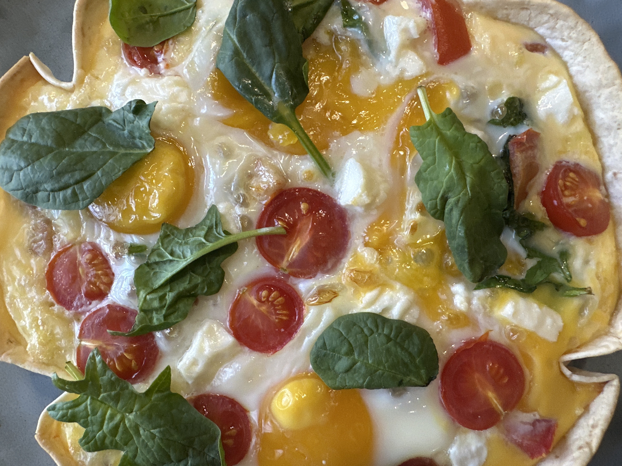 A close-up overhead shot reveals a tortilla bowl filled with cooked eggs, halved cherry tomatoes, spinach leaves, and small white cheese cubes. The egg yolks are visible and runny. The tortilla shell has a golden color. The scene is likely of food. There is no information to suggest the location is Chaikovsky, Russia.