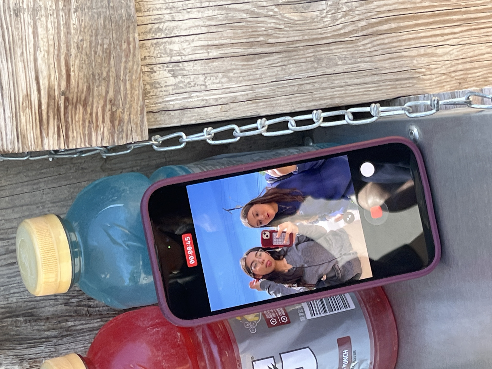 The image shows a close-up of a phone resting on top of a blue and a red sports drink bottle, which are themselves resting on a weathered wooden surface with a metal border and chain. The phone's screen displays a video of two young women taking a selfie, with one holding another phone. The lighting appears to be natural daylight. The location is unknown.