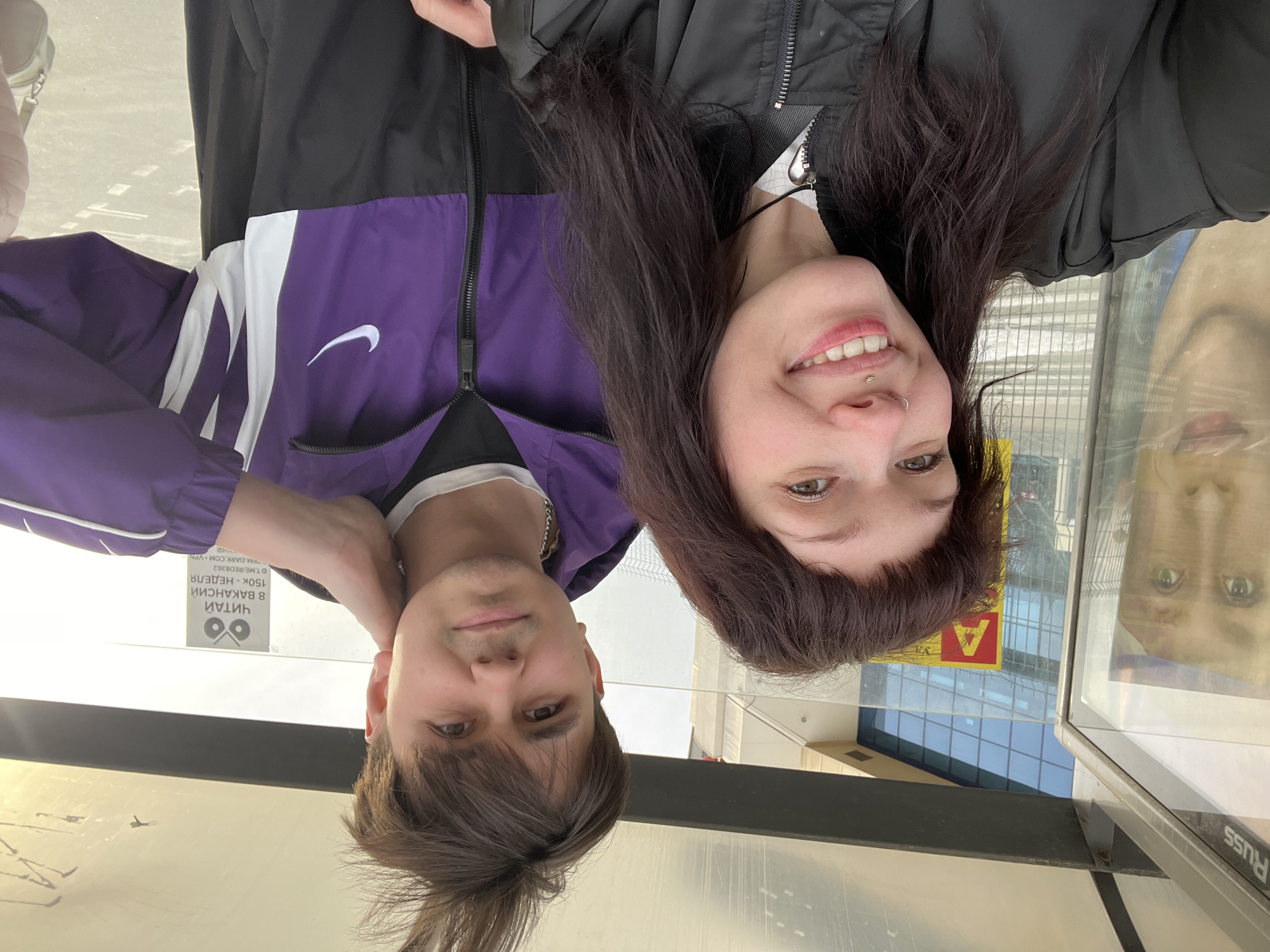 A photograph shows two people and their reflection on glass. A fair-skinned male with dark hair is wearing a purple and black Nike jacket and a chain. A fair-skinned female with dark hair is wearing a black jacket. The reflection in the glass shows another person with fair skin and dark hair. A sign in Russian is visible behind the people. The location is Kazan, Russia.