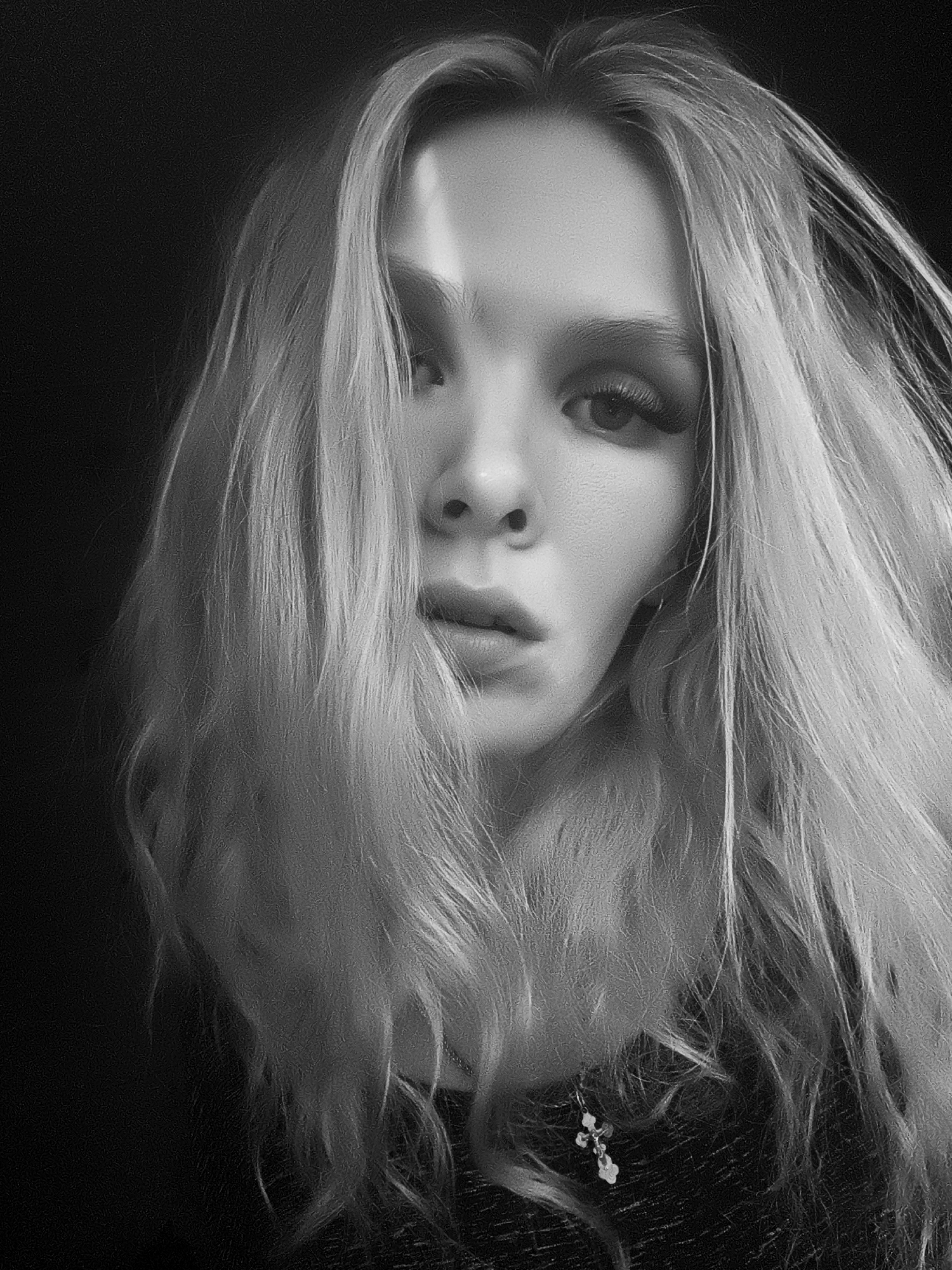 This is a black and white, close-up, portrait photograph of a young woman. She has long, blonde hair that is styled loosely and falls around her face, obscuring part of it. A sharp shadow cuts across her forehead and the bridge of her nose, creating a dramatic effect. Her eyes are dark and appear to be looking directly at the camera, with noticeable eyelashes. Her lips are slightly parted, giving her a contemplative or perhaps surprised expression. She is wearing a necklace with a small, ornate pendant that is visible in the lower right quadrant of the image. The background is dark and nondescript, providing a stark contrast to the subject. The lighting is dramatic, with strong highlights and shadows. There is no visible text in the image. The context provided places the image in Русский Вожой, Russia, but there are no specific visual cues within the photograph to confirm or deny this location. The overall mood of the image is somewhat mysterious and intense due to the lighting and the woman's direct gaze.