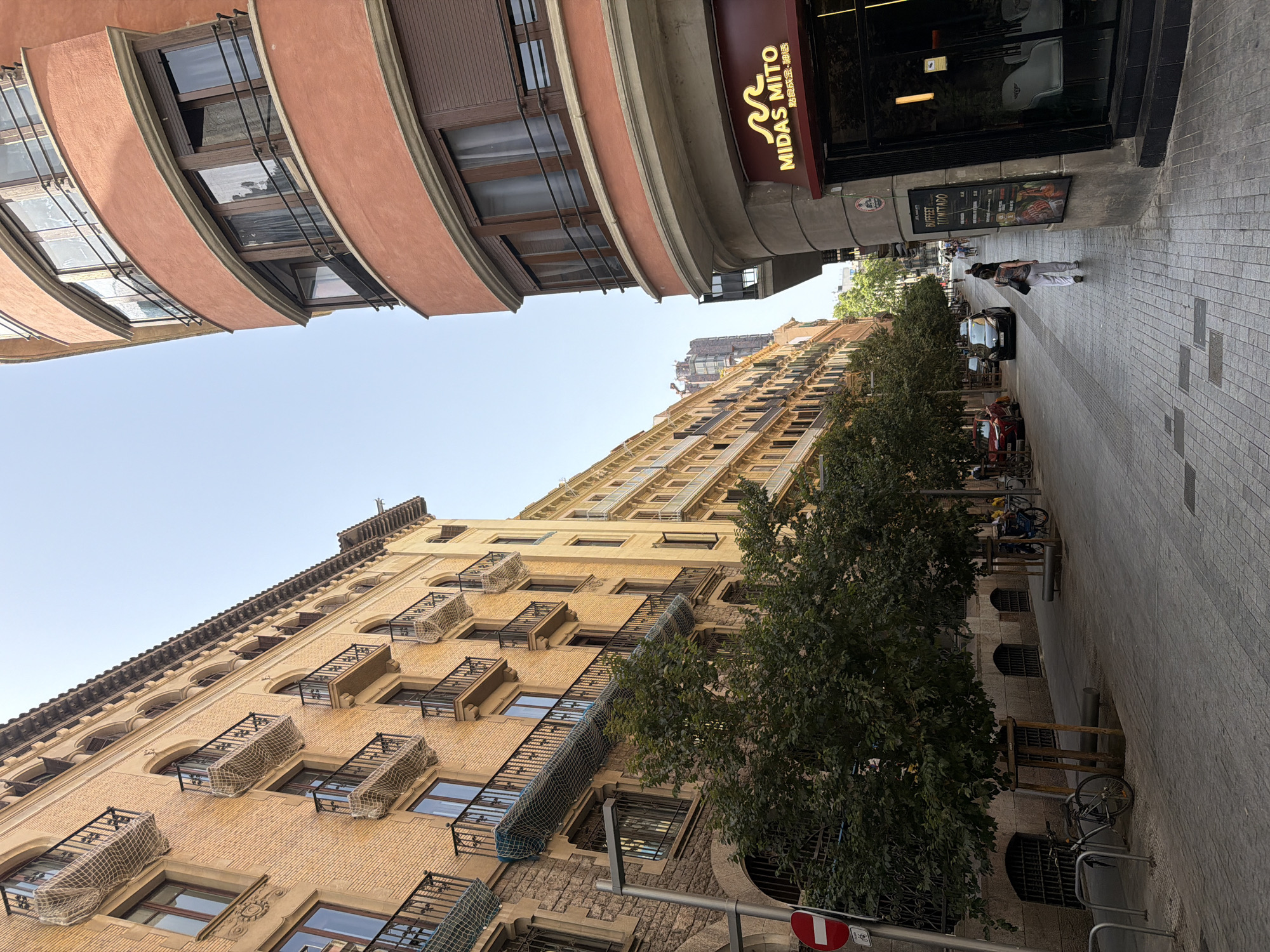 This is a ground-level, eye-level, medium shot of a street scene in Barcelona, Spain, likely in the Eixample district due to the architecture. The image captures a section of a pedestrianized street flanked by tall, multi-story buildings. On the left, a light yellow brick building features numerous balconies with black wrought-iron railings and protective netting, indicative of areas where construction or renovation may be occurring. The building has arched windows on the lower levels, some with barred openings. Directly in front of this building, several trees are planted along the sidewalk, some in wooden planters. A bicycle is parked near the curb. A circular red and white sign with a bicycle symbol is visible on a pole.
On the right side of the street, a curved building with a salmon-pink facade stands out. This building has multi-paned windows and dark brown shutters. At the ground level of this building, there is a storefront with a prominent sign that reads "MIDAS MITO" in a stylized font, below which are Chinese characters. A menu board is displayed outside the establishment.
The street itself is paved with cobblestones. A few people are walking along the street, including a woman in white pants and a patterned top walking away from the camera towards the right. A few cars are parked and driving on the street in the distance. The sky above is clear and light blue. The overall impression is of a typical urban streetscape during daytime in a European city.
