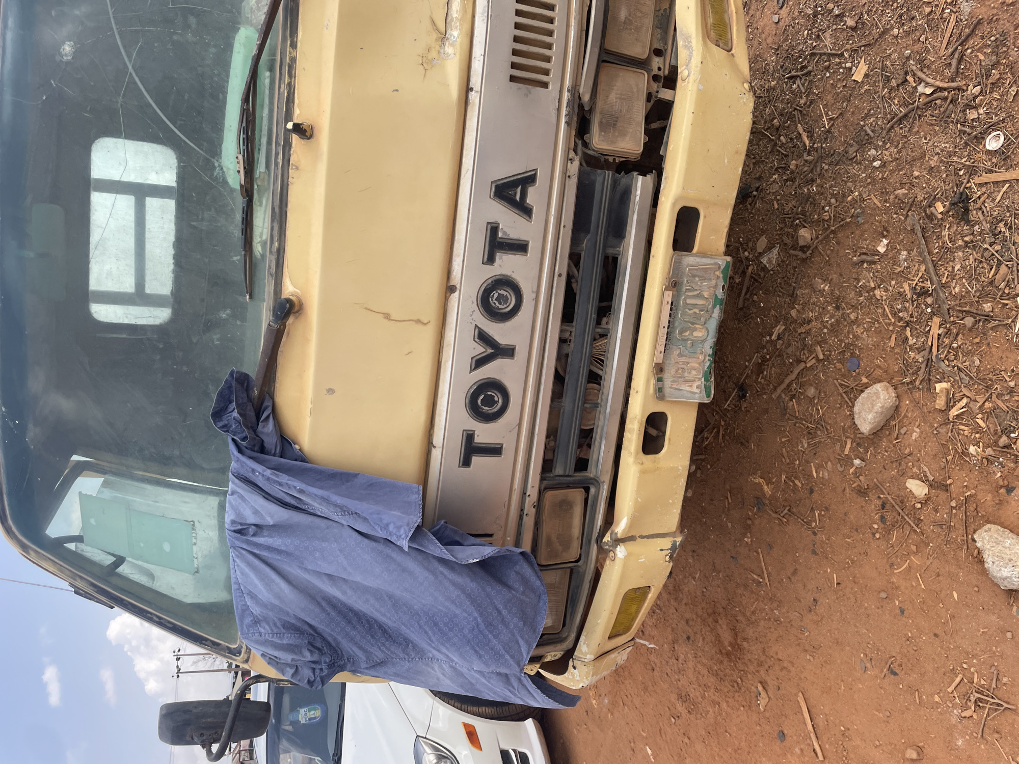 An old, light yellow or beige Toyota truck is the central subject, seen from a slightly elevated frontal perspective. The truck is parked outdoors on reddish-brown, dusty ground littered with sticks, small stones, and various debris. The clear blue sky with some white clouds is partially visible in the upper left.
The truck shows significant signs of age and wear. Its paint is faded and chipped, with visible dirt and minor damage on the bumper and body panels. The windshield is cracked and appears dirty, and a blue, patterned long-sleeved shirt is draped over its lower section and the hood. The headlights are dirty and appear to be old or damaged.
Prominently displayed vertically on the truck's grille is the text "TOYOTA". A license plate, predominantly green and white, is affixed to the front bumper on the right side. On the license plate, the letters "ABJ" are clearly visible, followed by "137" and possibly other faded characters like "EY". Part of a white car is visible behind and to the left of the truck's front bumper. The scene suggests a dry, outdoor setting, consistent with Galadimawa, Nigeria. No people or specific activity are visible.