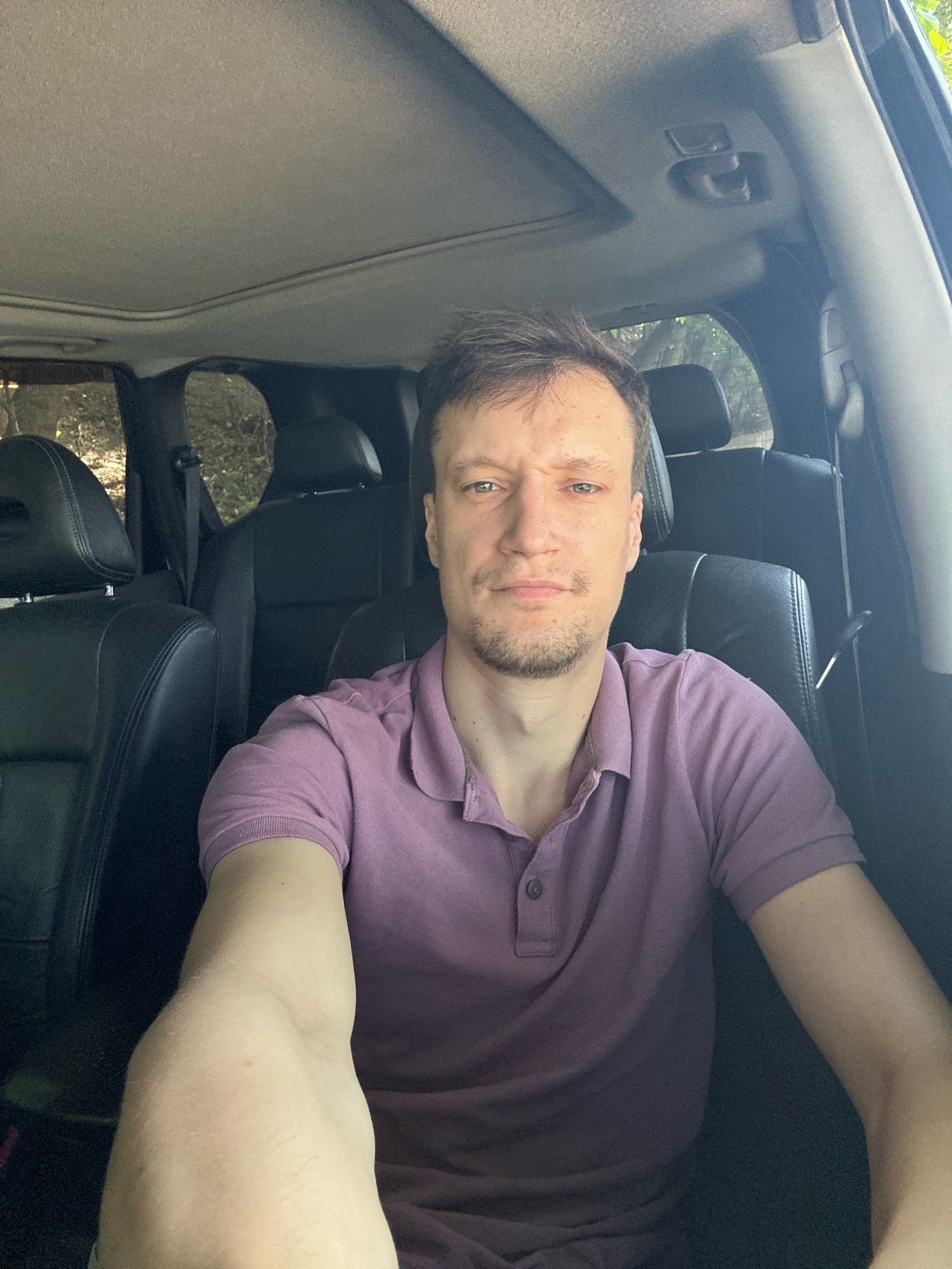 A young man with short brown hair, a light beard, and a purple polo shirt is sitting in the driver's seat of a car. He is looking directly at the camera with a neutral expression. The interior of the car is dark, with black leather seats visible. Sunlight is filtering in from outside, illuminating the man's face and arms. The location is not specified in Makiv, Ukraine.