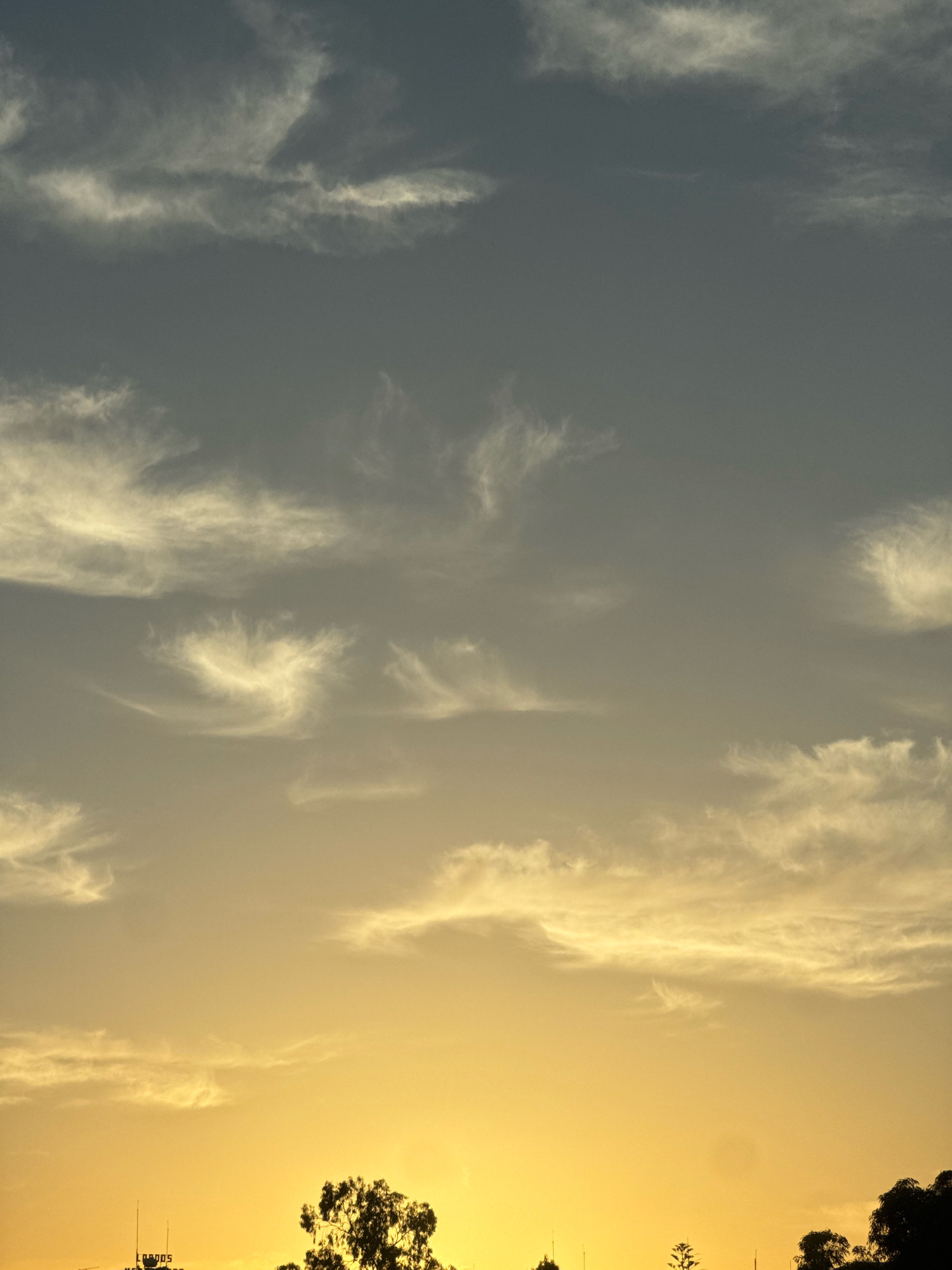 The image shows a dusky sky over the Agios Tychona Community in Cyprus. Wispy white clouds are scattered across a gradient of grey and yellow hues, indicating either sunrise or sunset. The bottom of the frame features a silhouetted treeline with a few dark green trees. Below the trees, a portion of a building with lettering is visible, along with two slender antennae. The scene is peaceful and devoid of any human activity.