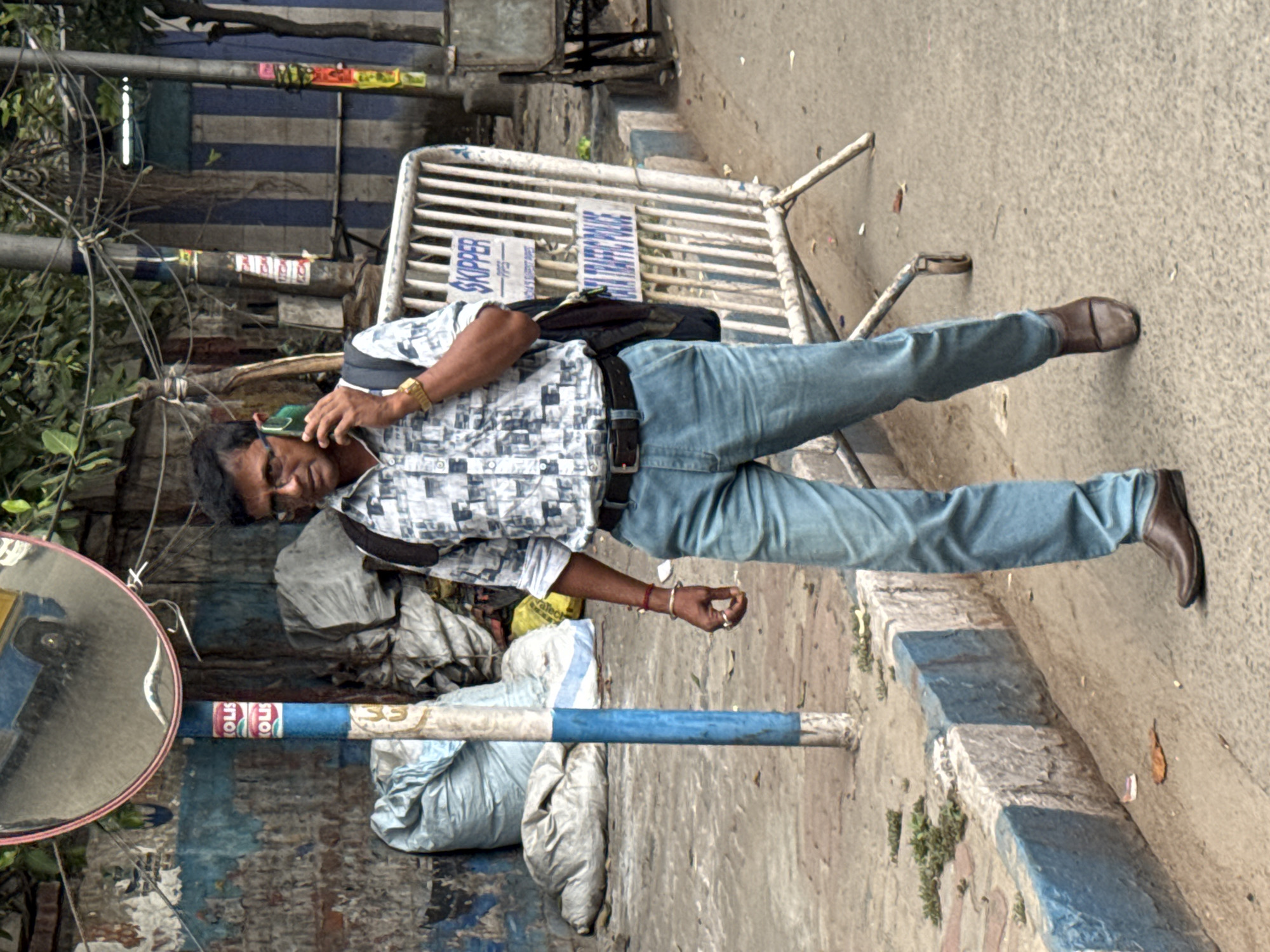 A man stands on a street in Kolkata, India, talking on a cell phone. He wears glasses, a patterned short-sleeved shirt, blue jeans, and brown leather shoes. A black bag is slung over his shoulder. He stands next to a blue and white striped curb. In the background, there's a white metal barrier, a utility pole with wires, and a building with blue and white stripes. There is also a round mirror affixed to a pole, with sacks and assorted objects piled nearby. The street surface is cracked and worn.
