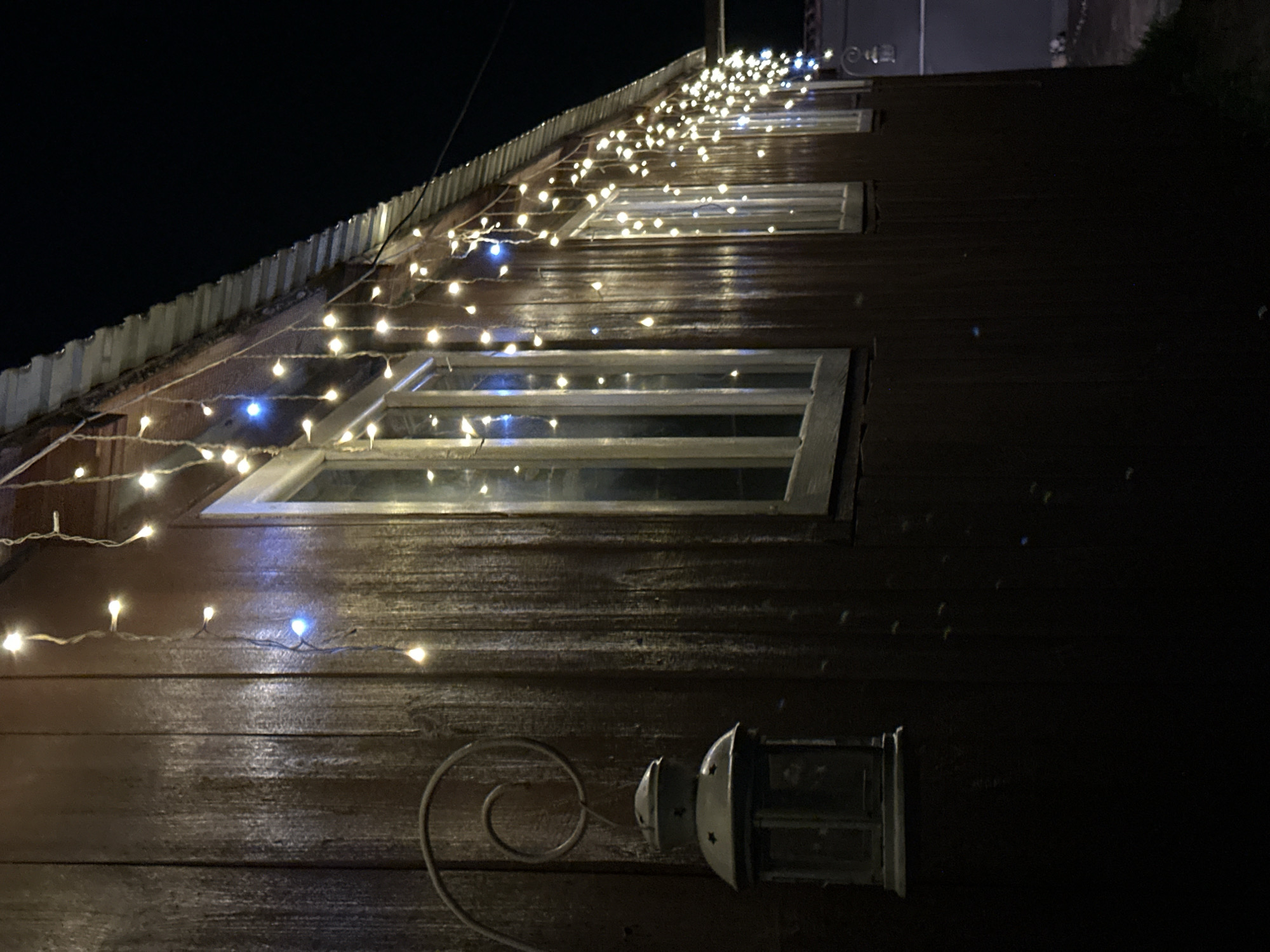 The image depicts the exterior of a wooden building at night, decorated with a string of fairy lights. The wooden walls are dark brown and appear to be made of vertical planks. A metal roof is visible along the top edge of the building. Several windows with white frames punctuate the facade. A decorative metal lantern hangs from a scrolled bracket on the left side of the building. The fairy lights, emitting a warm, yellowish glow with some blue accents, are draped along the roofline. The background is dark, indicating nighttime. The location is identified as Abzakovo, Russia.