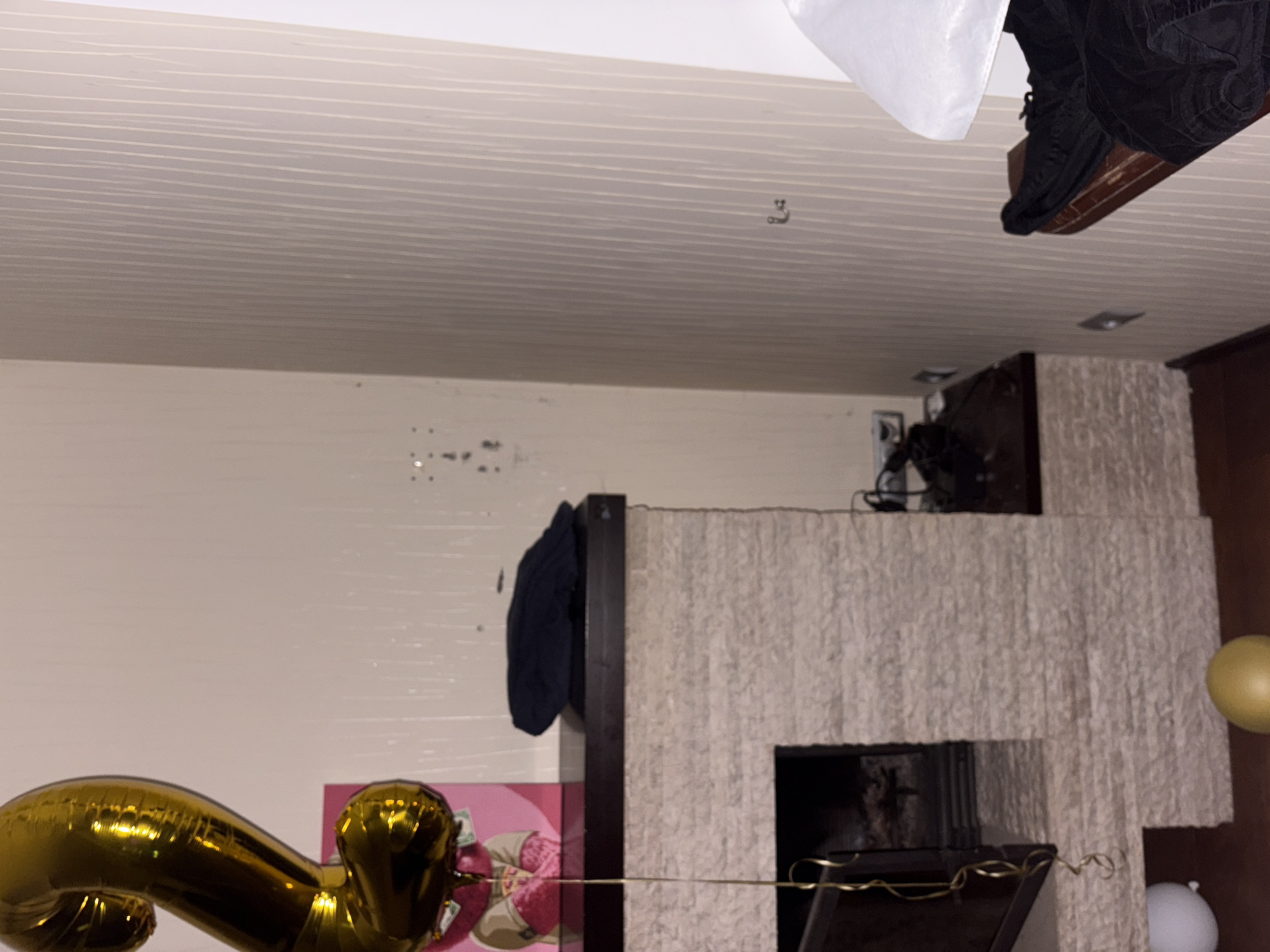 The image depicts an indoor scene, likely a living room, with a focus on a fireplace and surrounding decor. A large, gold, balloon shaped like the number "2" is prominently displayed to the left, hanging by a golden ribbon. To the right of the balloon, a pink framed picture featuring a teddy bear is visible. The fireplace itself is constructed with light-colored, textured stone. A dark, rectangular object, possibly a piece of clothing, rests on the dark mantelpiece above the fireplace.
The walls in the background are covered with wallpaper. The left wall has a light, neutral tone with some scattered dark marks. The right wall is adorned with vertical stripes in a similar light shade.
In the foreground, to the left of the fireplace, a white balloon is partially visible on the floor. To the right of the fireplace, a golden, spherical object, possibly another balloon or ornament, rests on the floor.
Further to the right, a wooden surface is visible, with dark fabric draped over it. Above this surface, a portion of a white lampshade can be seen.
The overall impression is that of a decorated interior, possibly for a celebration. The image does not contain any elements that would allow for the identification of a specific location within the city of Orekhovka, Russia.