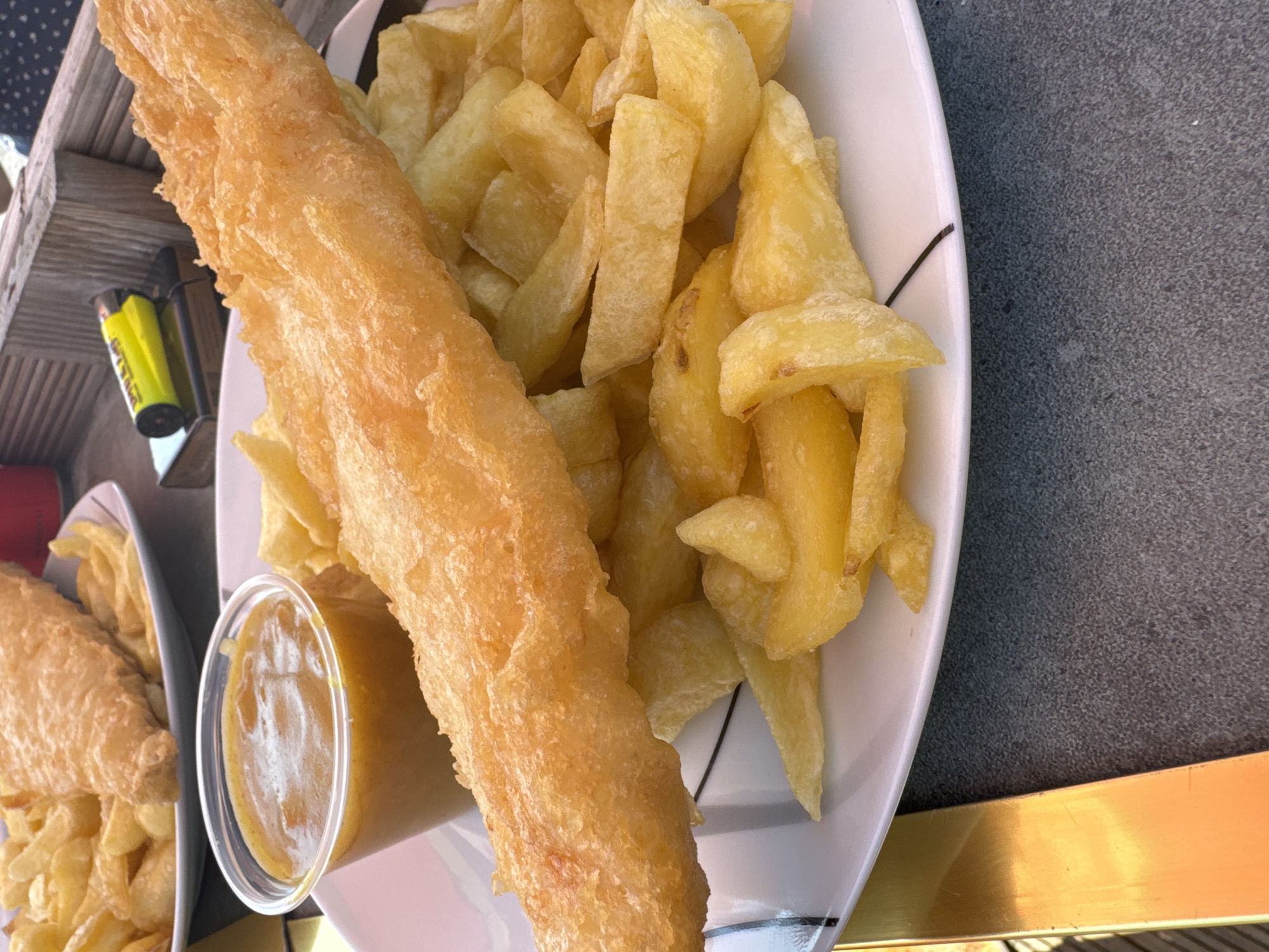 This image captures a close-up view of a meal of fish and chips, likely served in Great Yarmouth, United Kingdom. The primary subjects are a large piece of battered fish and a generous portion of thick-cut chips, presented on a white plate with a subtle black line design along the rim. A small, clear plastic container filled with a yellowish sauce, possibly curry sauce, sits to the left of the fish. Another plate of fish and chips is partially visible in the background to the left.
The food appears freshly prepared, with the batter on the fish golden brown and slightly crispy, and the chips uniformly cut and cooked. The setting suggests an outdoor dining environment, with a wooden surface visible on the left and a textured, grey surface on the right, possibly a table. A yellow lighter is also visible on the wooden surface, hinting at a casual meal or an outdoor setting where one might be used.
The lighting in the image suggests it was taken during daylight hours, likely late morning or afternoon, given the brightness and shadows. There are no people visible in the frame, so any emotions or specific activities beyond eating are inferred. The overall impression is one of a classic British seaside meal. No visible text is present on the food itself or the plates.