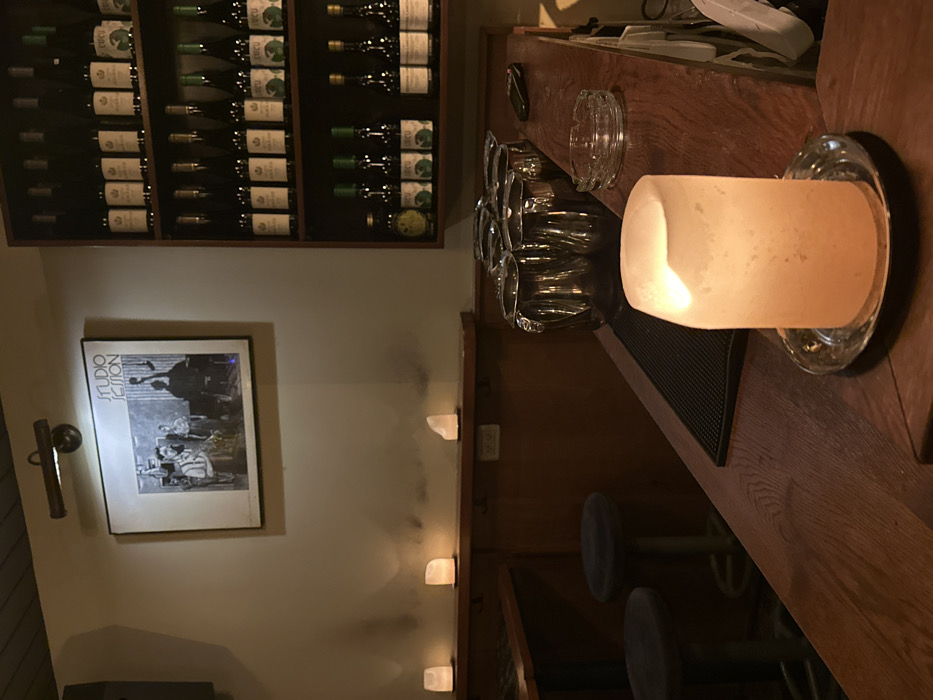The image displays the interior of an establishment, likely a bar, characterized by dim ambient lighting. A prominent dark wooden bar counter extends from the foreground towards the midground on the right. In the immediate foreground, centered on the counter, a glowing, translucent, block-shaped lamp, possibly made of frosted glass or salt crystal, rests on a circular silver-toned coaster. Behind this lamp, a black rubber bar mat is visible, followed by a row of six polished metallic pitchers. Further along the counter are a clear glass ashtray with multiple cigarette rests and a black remote control.
To the right, a dark wooden shelving unit is affixed to the wall, holding multiple rows of identical bottles. The top two shelves contain bottles with light-colored labels and dark caps. The middle shelf displays bottles predominantly with green labels, some marked "IBIU," and one bottle with a yellow label. The bottom shelf presents a mix of green-labeled and light-labeled bottles.
On the left side of the frame, a light-colored wall features a framed black-and-white photograph titled "STUDIO SESSION," depicting a group of musicians, including one with an upright bass. Above the frame, a brass-colored spotlight illuminates the artwork. Below the photograph, on a narrow wooden ledge, three additional glowing, translucent, block-shaped lamps are evenly spaced. Two dark-upholstered bar stools are partially visible at a dark wooden table section in the lower-left. A dark speaker is present in the upper-left corner of the wall. No individuals are depicted. The precise location within Tel-Aviv, Israel, is not identifiable from the visual content.