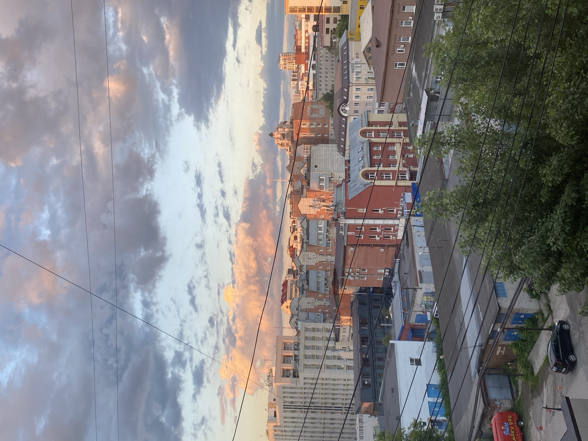 The image depicts a cityscape in the city of Perm, Russia, under a cloudy sky tinged with the warm colors of sunset. In the foreground, power lines crisscross the frame. Below the sky, a dense collection of buildings forms the urban landscape. Closer to the bottom of the frame, there are low-lying industrial or garage-like structures, some with blue doors and graffiti. A black car is parked on a paved area next to one of these buildings, and a red food truck labeled "SMOKY DOG" is also visible. Further back, a mix of residential and commercial buildings, varying in height and architectural style, rise up. These include multi-story buildings with light-colored facades, red brick structures with numerous windows, and some with rounded rooflines. The overall scene captures a slice of urban life in Perm at dusk, with a contrast between the functional lower buildings and the more prominent structures behind them, all under a dramatic sky.