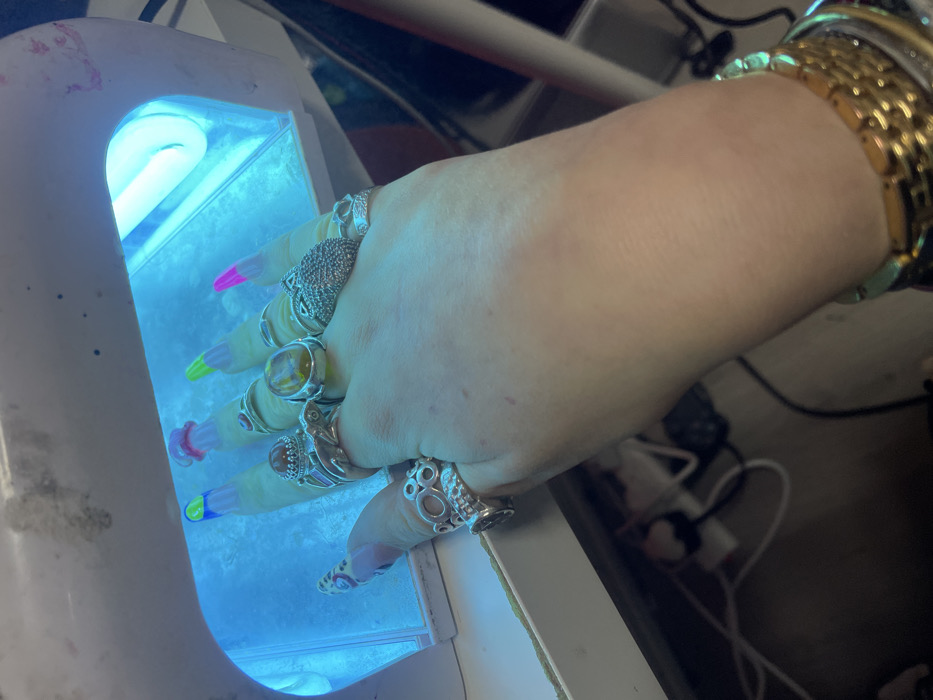 A hand adorned with multiple rings, including one with an amber stone, is placed inside a UV nail lamp. The nails are painted with different bright colors on the tips. Gold bracelets are on the wrist. Cords and plugs are visible in the blurred background.