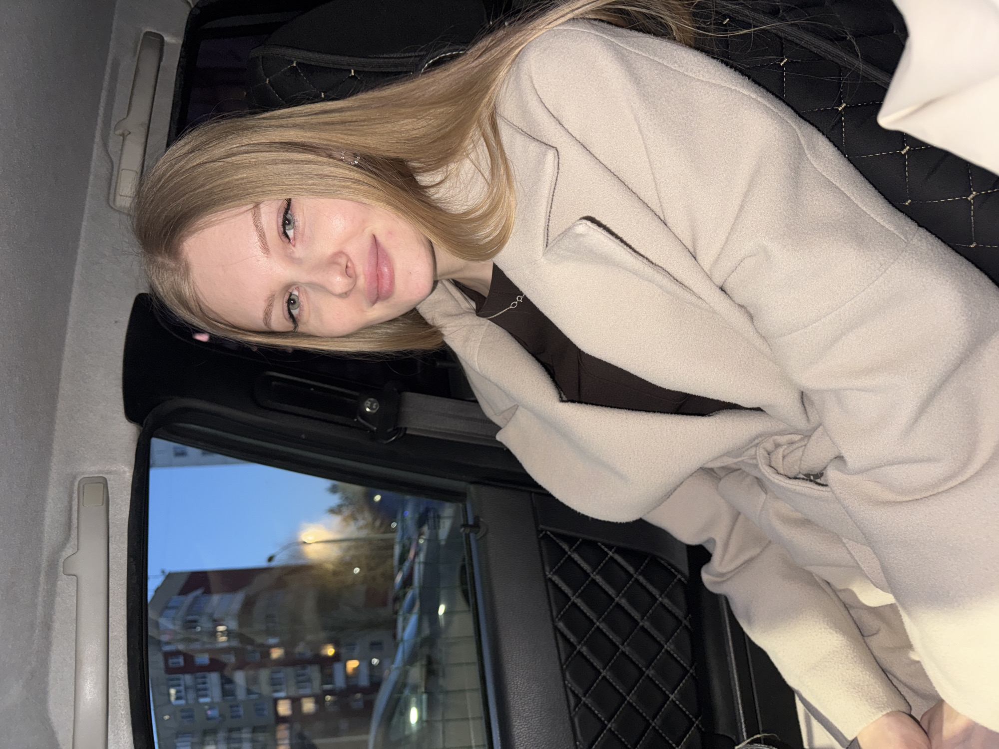 A fair-skinned woman with long, blonde hair is sitting inside a vehicle, possibly a car, as suggested by the side window visible. She is wearing a light-colored jacket and a dark shirt underneath. The background outside the window reveals a street scene at dusk in the city of Perm, Russia. Buildings, streetlights, and the darkening sky are visible. The interior of the vehicle features a black quilted seat and a white ceiling.
