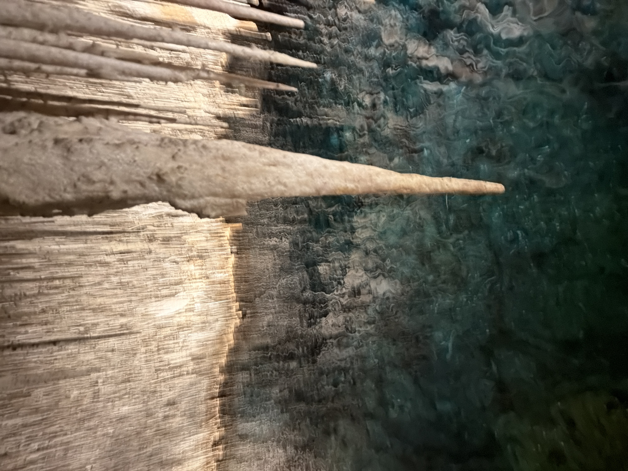 The image depicts a close-up view of stalactites inside the Caves of Drach, located in Manacor, Spain. The stalactites are light-colored and hang from the cave ceiling. The background shows a body of water, likely an underground lake, with a rippled surface reflecting the cave's interior. The water appears dark green and blue due to the lighting and depth.