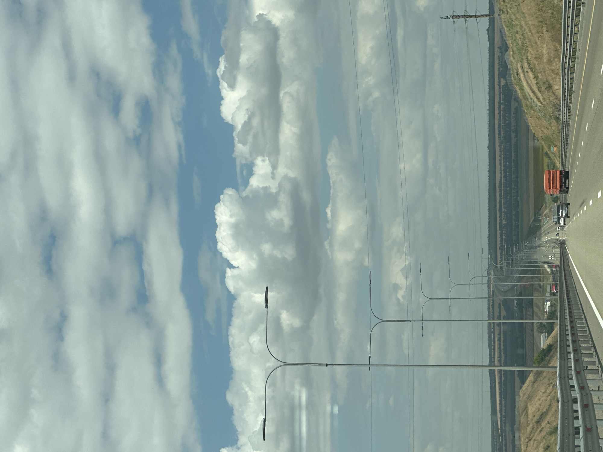 The image depicts a view from a highway in the vicinity of Grushevskaya, Russia, on a cloudy day. The highway stretches into the distance, with multiple lanes and a metal guardrail on the right side. Tall lampposts line the left side of the road. In the background, rolling hills covered with dry grass and scattered vegetation are visible, along with a band of trees and agricultural fields. Several vehicles, including cars and a large orange truck, are driving on the highway. Power lines are strung across the sky. The sky is filled with a mix of white and gray clouds.