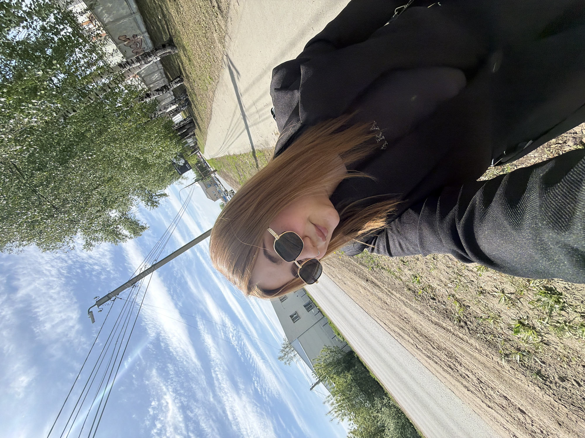 A woman in sunglasses and a dark jacket is outdoors. The photo appears to be a selfie, taken on a sidewalk next to a road. A building is visible in the background. The sky is blue with streaky clouds. A utility pole with wires is on the left. Trees line the road. Based on the context clues and the presence of birch trees, the location is in Chaikovsky, Russia.