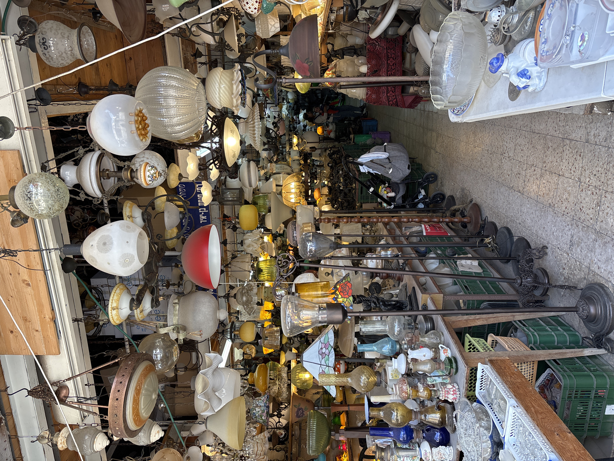 The image depicts a densely packed alley in Tel Aviv, Israel, likely a marketplace or antique district. The space is filled with a vast collection of vintage light fixtures, primarily chandeliers and standing lamps, hanging from above and displayed on shelves or the ground. The light fixtures vary in style, size, and material, including glass, metal, and ceramic components. The alleyway is narrow, with the fixtures creating an almost claustrophobic effect. The floor is tiled, and there are some visible containers (baskets and plastic crates) used for storage or display. There are a few people visible in the distance, browsing the items. There is a baby stroller in the aisle on the right. The lighting is a mix of natural daylight and the ambient glow of the fixtures themselves.