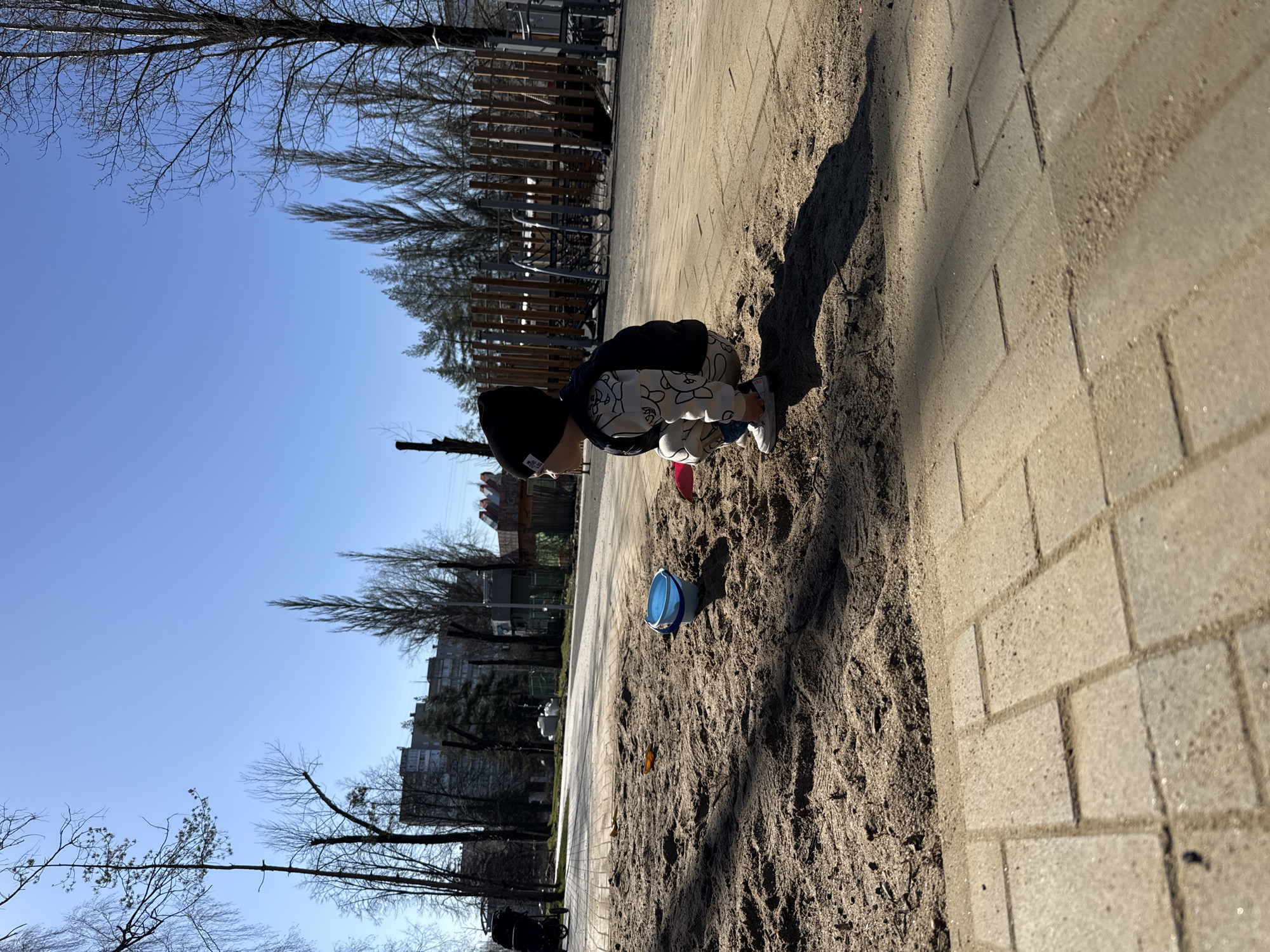 A child wearing a black beanie, patterned white pants, and a dark jacket is crouched down in a sandbox. The sandbox is situated next to a paved area with brickwork. A blue bucket and a red object are in the sand nearby. The setting appears to be a playground or park, possibly in the Rusanovo district of Russia, considering the query. Buildings and bare trees are visible in the background under a clear blue sky. A fence or playground structure is also in the background.