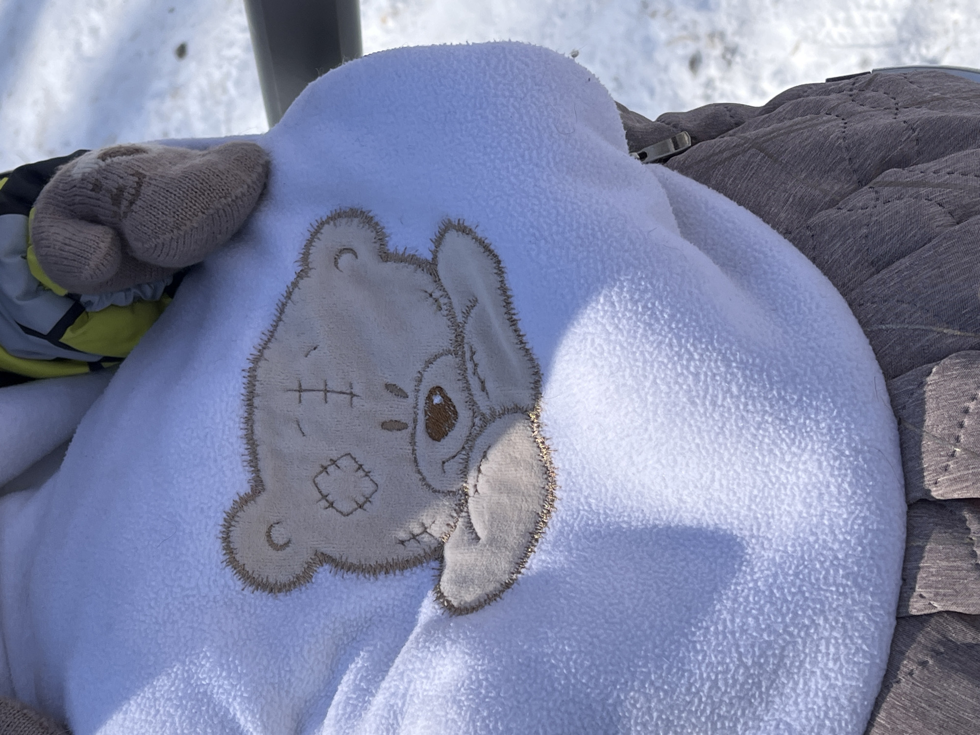 A close-up shot reveals a white fleece baby snowsuit with a stitched teddy bear design on its front. A knitted mitten lies atop the snowsuit, and a quilted grey fabric is visible in the background. The presence of snow in the background, visible above the snowsuit, suggests the image was taken in a cold environment, consistent with the climate of Krasnoyarsk, Russia.