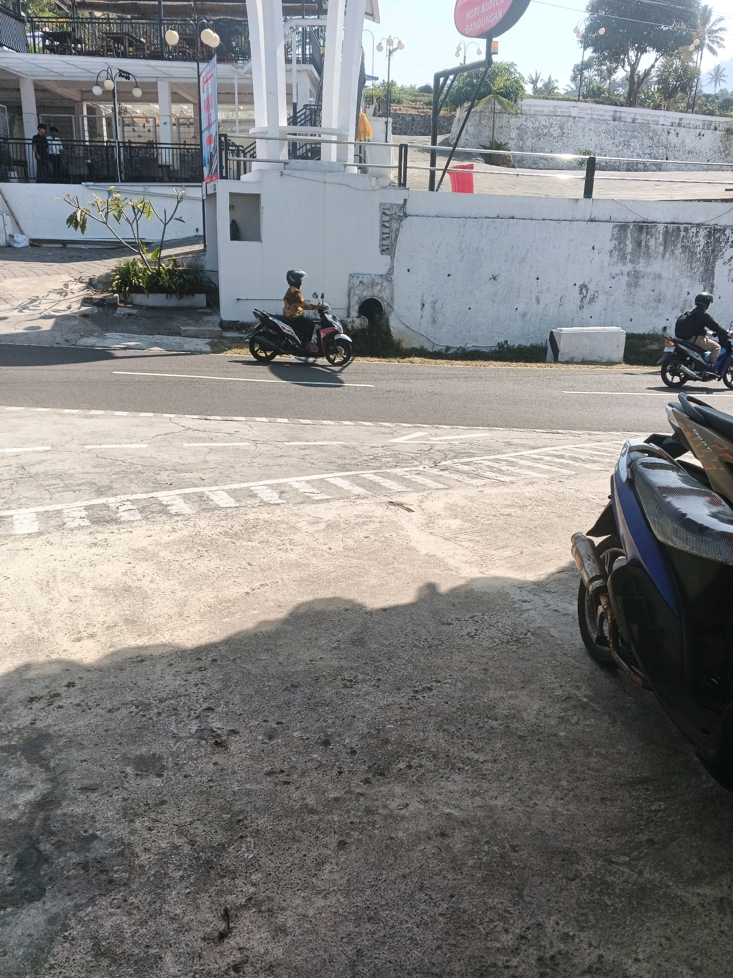 In Pakopen, Indonesia, a scene unfolds on a street with a concrete and asphalt surface. Two individuals are on motorcycles. One person, wearing a helmet and a patterned shirt, rides a pink and white motorcycle from left to right across the frame. Another individual, also helmeted and with a backpack, rides a dark-colored motorcycle from right to left. In the background, a white building with multiple levels features a balcony with tables and chairs. A red trash bin is visible near the building. The sky is clear and blue.