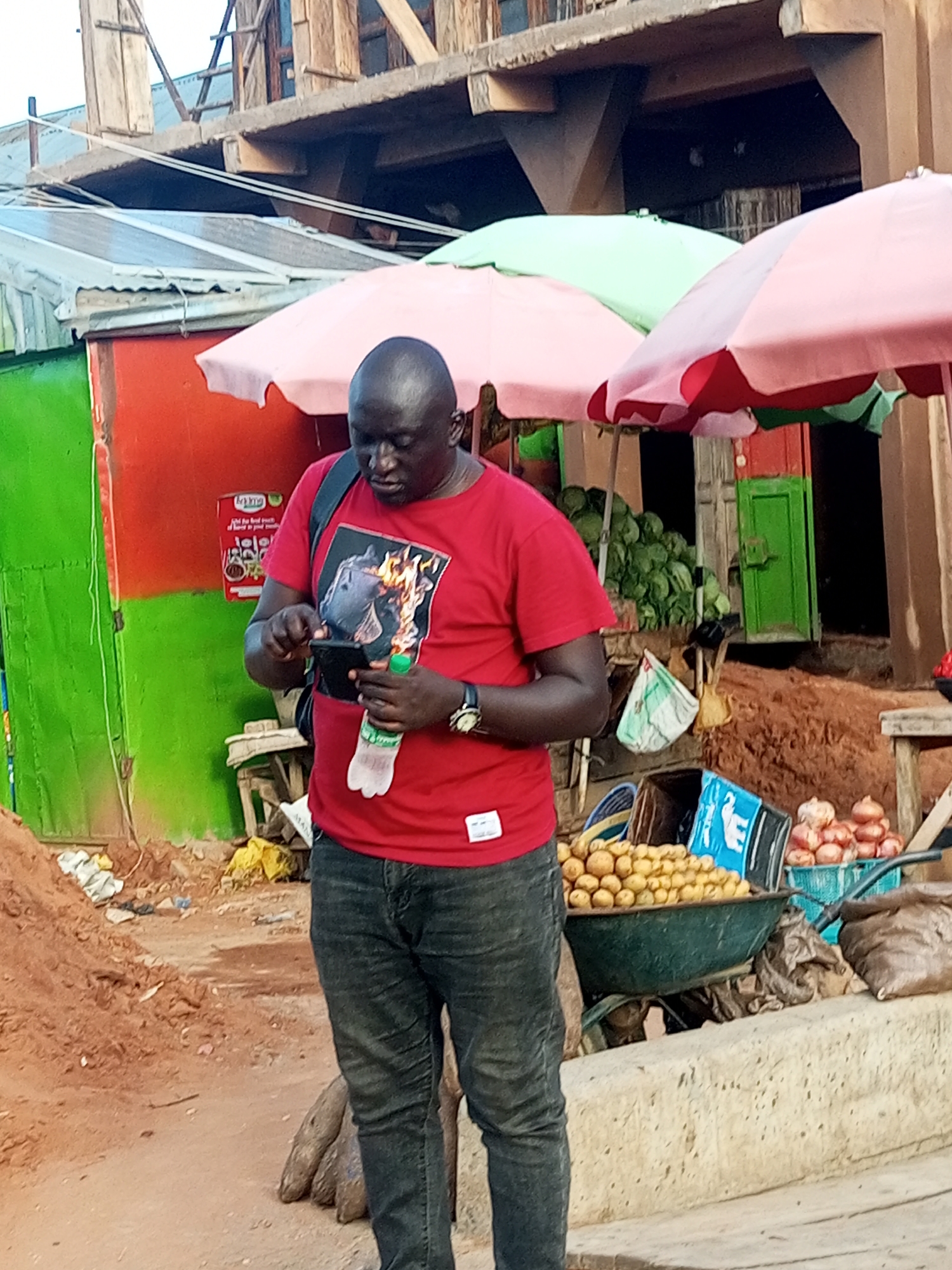 An overhead shot captures a Black man in a red t-shirt and black jeans standing in a market setting. He holds a smartphone and a bottle of water. Behind him, a market stall displays fresh produce, including a wheelbarrow full of potatoes and a stack of onions. The background reveals a building under construction, with wooden beams and concrete elements. The market appears to be located in Gombe, Nigeria, evidenced by the surroundings and the general atmosphere of an open-air marketplace. The man is engaged with his phone, suggesting he is either making a call or browsing the internet. The overall scene depicts a moment of everyday life in a Nigerian marketplace.