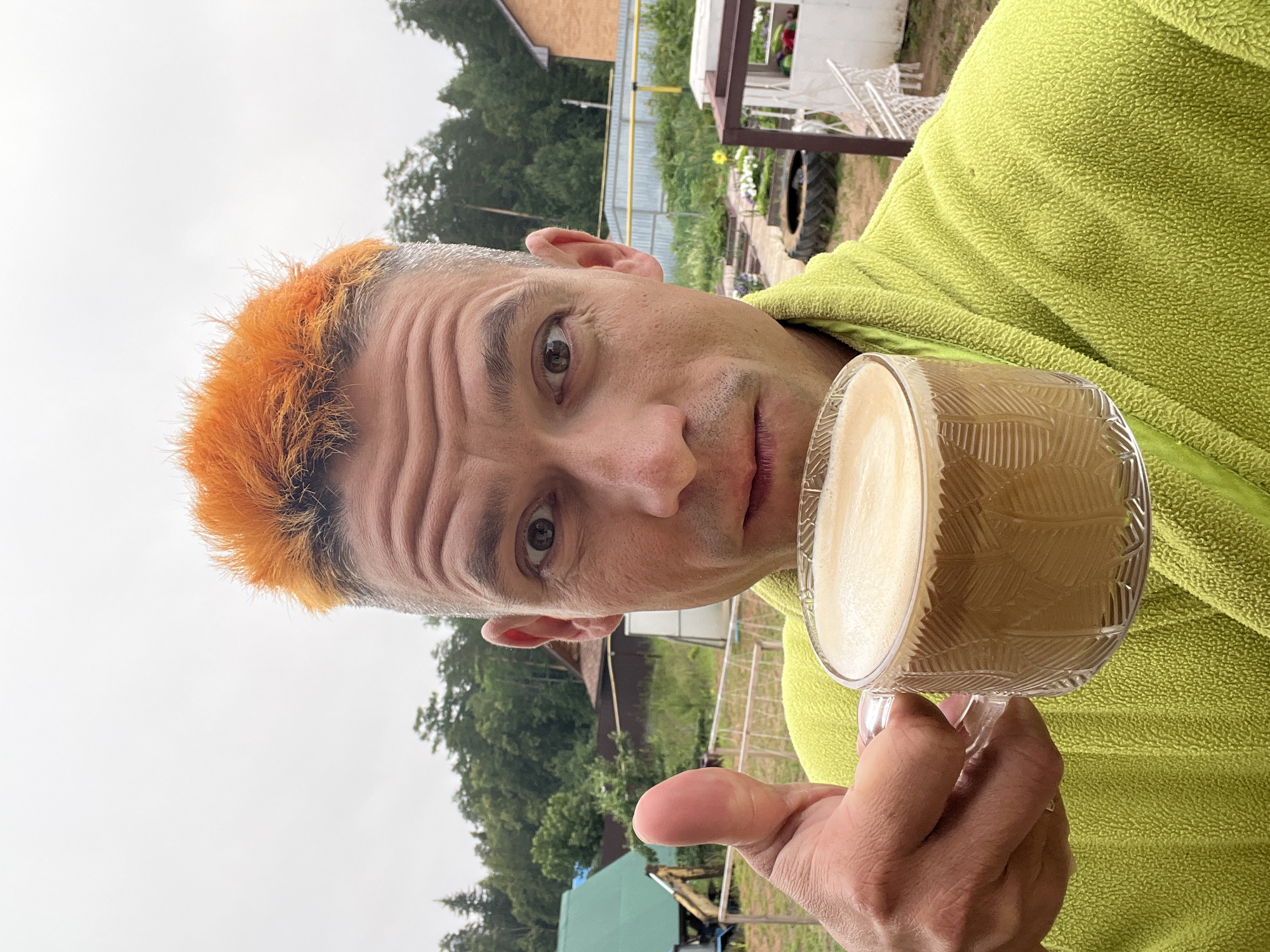 A man with bright orange, spiky hair and a light stubble on his chin, wearing a lime green textured robe, holds a clear glass mug filled with coffee to the left side of his face. He is looking directly at the camera with a surprised expression, his eyes wide open. His right thumb is raised in a "thumbs up" gesture, positioned in the lower left quadrant of the image. The background appears to be a semi-rural or suburban setting, possibly in Chaikovsky, Russia, featuring a mix of green trees, a weathered wooden fence, and what looks like a garden area with raised beds and some white garden furniture. The sky is overcast and pale.