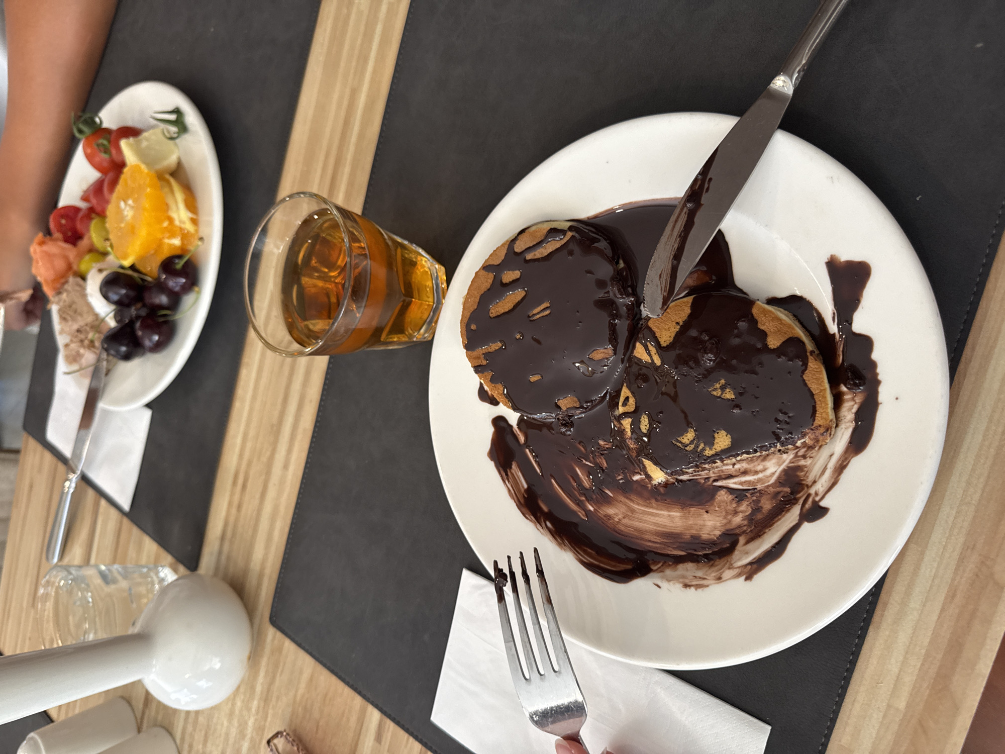 The image captures a close-up view of a white plate holding two chocolate-covered pancakes, with a knife resting on the edge of the plate. A fork, with some chocolate on its tines, is also visible on the left side of the plate, held by a hand not fully in frame. To the right of the plate, a short glass contains a amber-colored liquid. In the background, another plate with fruit and what appears to be some kind of salad is visible. The table is made of wood, with a dark placemat under the plates. A white ceramic object, possibly a creamer or sugar dispenser, is also present on the table. The setting appears to be a restaurant or dining area. The image was taken on a wooden table with a dark placemat, suggesting an indoor dining environment. The presence of two plates with food and drinks implies at least two individuals are dining. The food on the main plate consists of pancakes covered in chocolate sauce, while the background plate has a variety of fruits like cherries, oranges, and tomatoes, along with some leafy greens. The overall scene suggests a meal, possibly breakfast or brunch. The image's detail and composition suggest it was taken at a table in a restaurant or cafe. The details of the table setting, including the placemat and cutlery, contribute to the visual narrative of a dining experience.