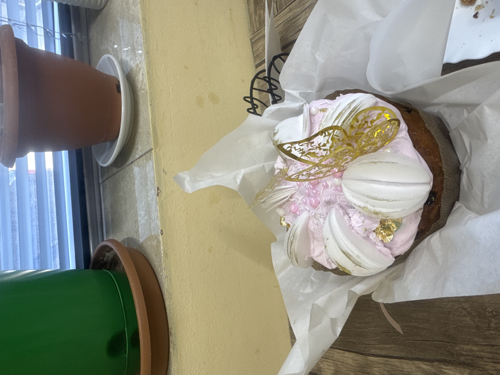 The image shows a decorated cake inside a white paper wrapper, likely a kulich, decorated with pink frosting, white meringue, and a gold butterfly. The cake is on a wooden surface next to a wall with two potted plants. The location is possibly in Tbilisi, Georgia, given the potential presence of a traditional kulich.