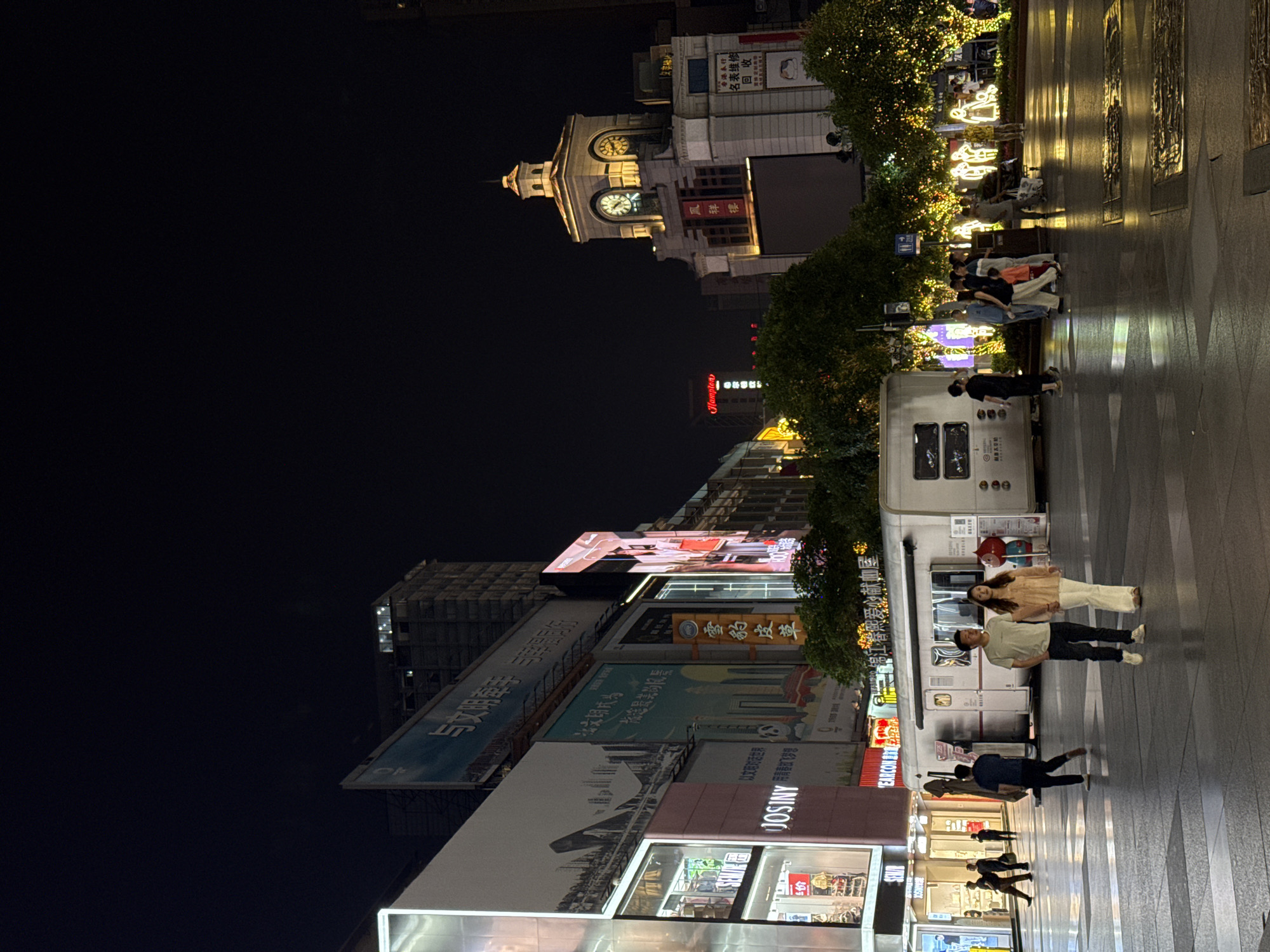 Here is a detailed description of the image:
The image presents a nighttime street scene in Jinjiang District, China, characterized by a blend of urban elements and illuminated signs. Pedestrians traverse a paved area in the foreground.
Notable buildings are present: a building with a prominent clock tower, illuminated trees and glowing decorations, and shops with bright signs and advertisements in Chinese characters. Brand names such as "JOSINY", "CARE ANISOC", and "Hamplon" are visible. Several billboards display various promotional content.
The individuals in the scene are walking or standing, and the scene appears to be lit by artificial lights and signs.