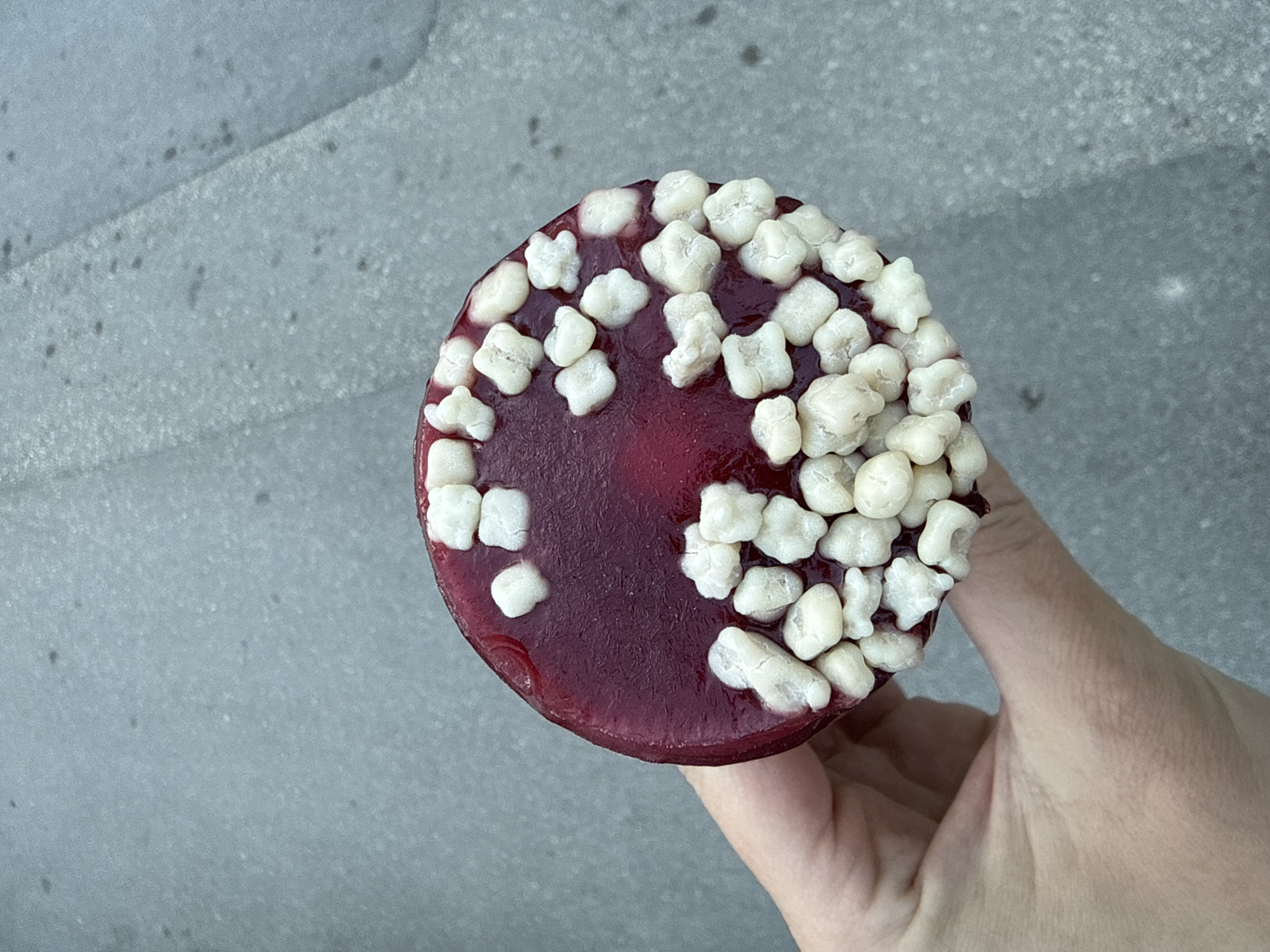 A person is holding a circular, reddish-purple dessert topped with irregularly shaped white pieces. The dessert appears to be a small cake or tart, possibly with a gelatinous topping. The white pieces are scattered across the surface. The person's hand is visible, with light skin tone. The background is a grey, textured surface, potentially concrete or stone. The location is not discernible from the visual data, and therefore it is not possible to confirm the location as Đenovići, Montenegro.