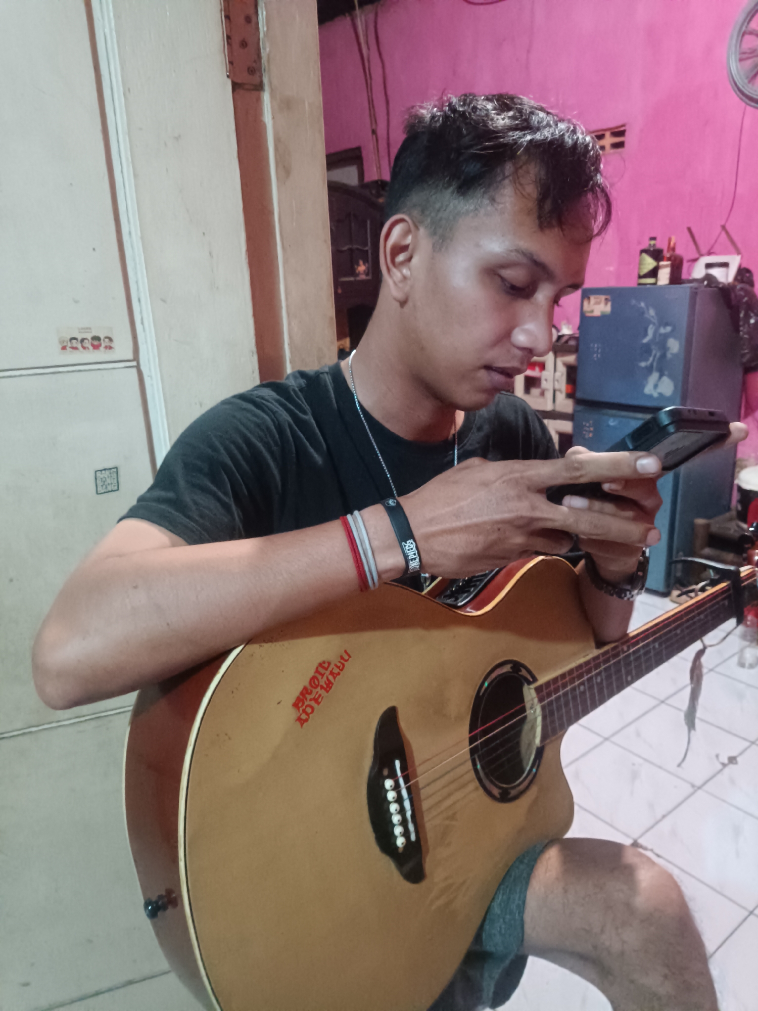 A medium shot shows a young Indonesian male with short, dark, styled hair, wearing a black t-shirt, and grey shorts, holding an acoustic guitar and a smartphone. He is seated in what appears to be a residential interior, possibly a room in Depok, Indonesia. The man is looking down at his smartphone, which he is holding in his right hand, while his left arm rests on the guitar. The guitar is a light brown acoustic with a dark soundhole and bridge. Red text is visible on the guitar's body, reading "BROIL ADEWANJU". The background is dimly lit with a pink wall on the right, a blue refrigerator, and other indistinct objects, suggesting a casual, lived-in space. The floor is tiled with a white and grey pattern. The overall impression is a candid moment of a person engaged with technology and musical equipment at home.