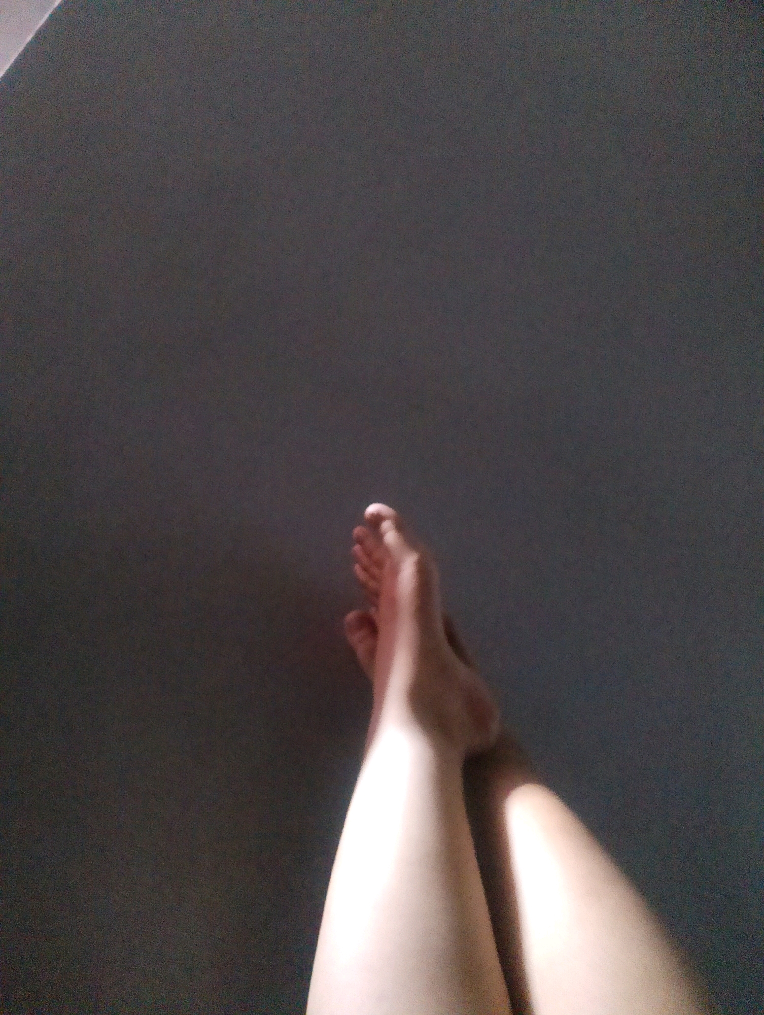 The image shows a close-up view of a person's legs and feet, illuminated by a diagonal beam of light that casts shadows on a dark, textured wall. The legs are bare, and the feet are positioned in a way that suggests they are resting or crossed. The lighting creates a dramatic contrast, highlighting the contours of the skin. The background appears to be an interior wall with a slightly rough texture. There is no indication of the city of Magelang, Indonesia, within the image itself.