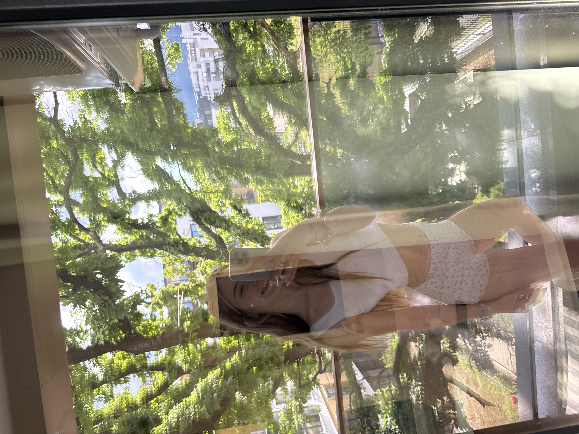 A woman takes a selfie in front of a window in Sofia, Bulgaria. Her reflection is superimposed over a view of trees and a building in the city. She wears a white top and patterned shorts. The window frame is visible, as well as an air conditioning unit on the left side of the frame.
