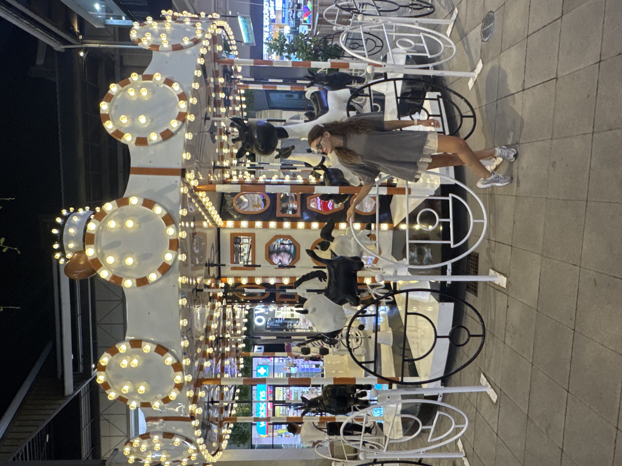 A woman stands next to a decorative carousel with cow figures in Baan Chaweng, Thailand. The carousel is adorned with illuminated circles. The woman wears glasses, a gray dress, and sneakers. The background includes storefronts with signs. The ground is paved with tiles.